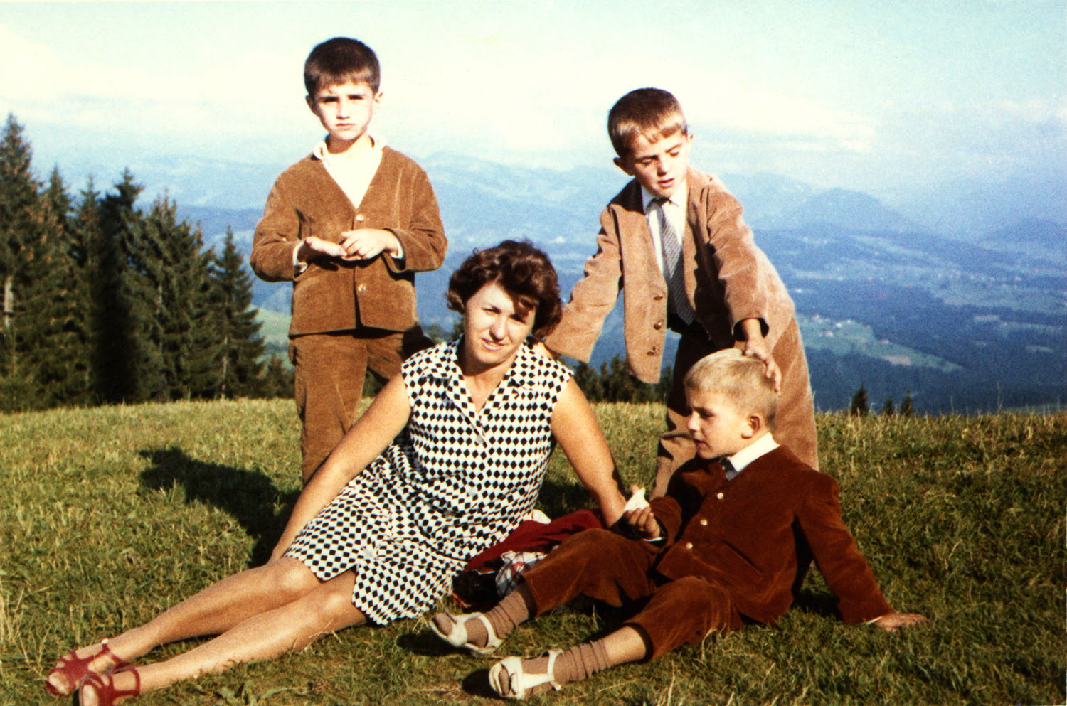 Une femme est assise dans l’herbe. Un jeune garçon est assis à ses côtés. Deux garçons sont debout derrière. On voit les montagnes en arrière-plan