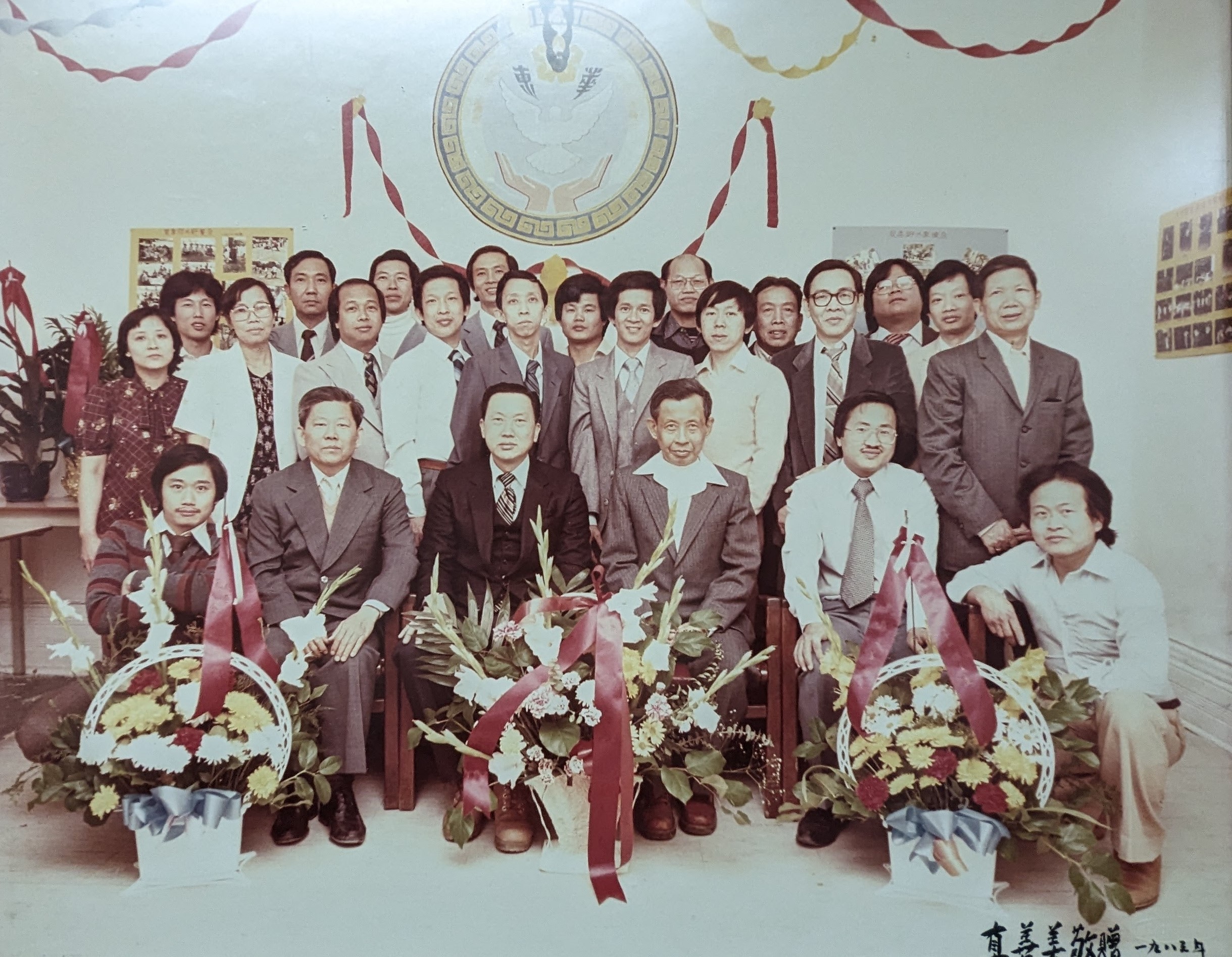 hoto couleur un peu délavée montrant un groupe de 24 personnes d’origine sino-cambodgienne réparties sur deux rangées prenant la pose de façon officielle, avec trois grands paniers de fleurs devant eux.