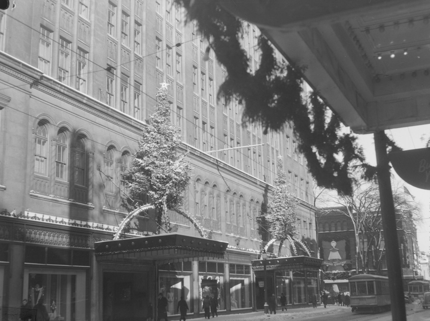 Sur la façade de la rue Sainte-Catherine, les marquises du magasin Eaton sont en 1946 décorées de grands sapins et d’arcs lumineux. Des passants circulent devant les vitrines, alors qu’un tramway passe.