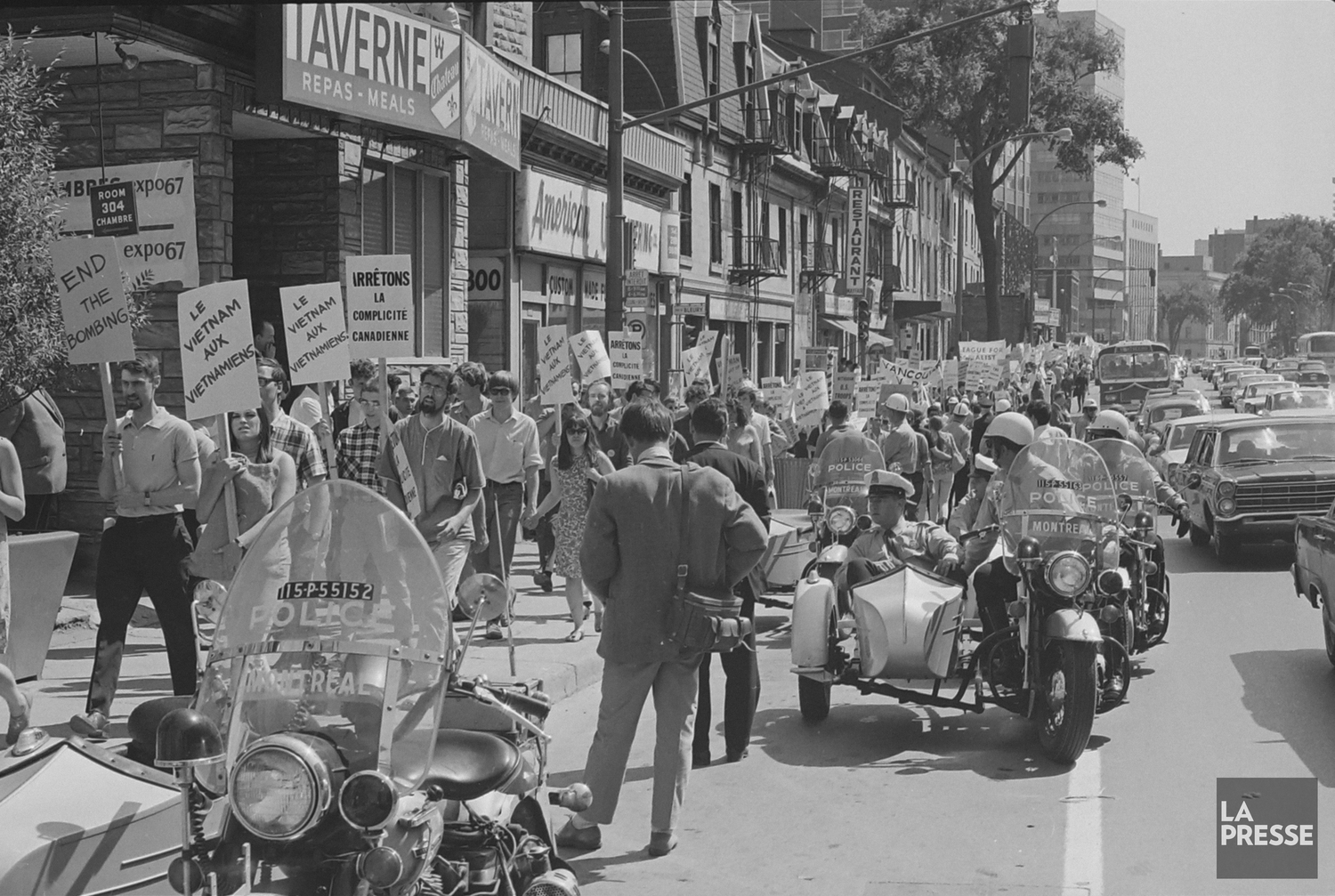 Manifestation au centre-ville, beaucoup de personnes ont des pancartes « Le Vietnam aux Vietnamiens » et autres messages anti-guerre. Des policiers à moto sont à l’avant-plan le long de la foule. 