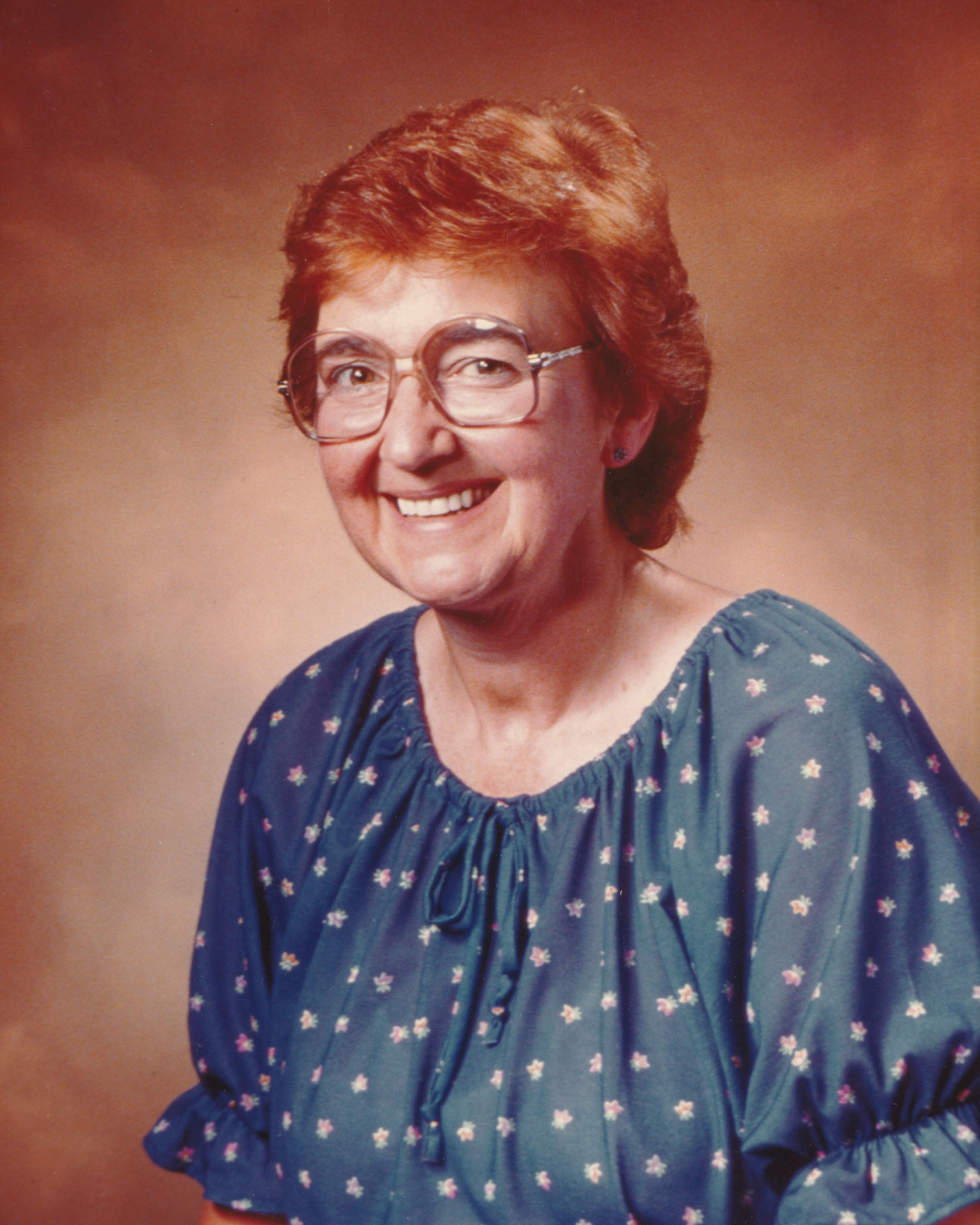 Une femme souriante portant des lunettes et un haut bleu avec des fleurs. 