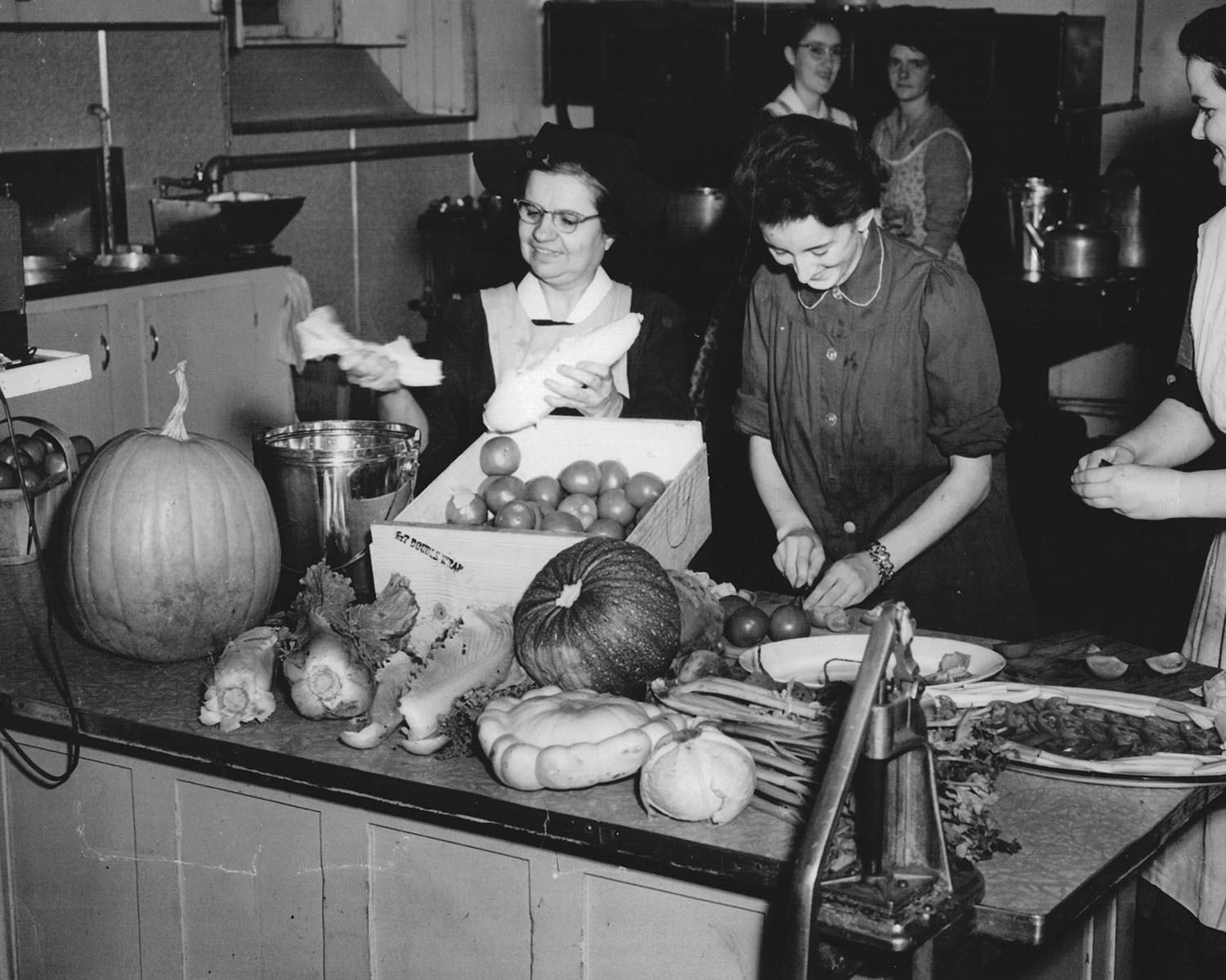 Deux femmes préparent des légumes sur un comptoir rempli d’une abondance de légumes. Derrière et à côté, trois personnes. Tous semblent s’amuser. 