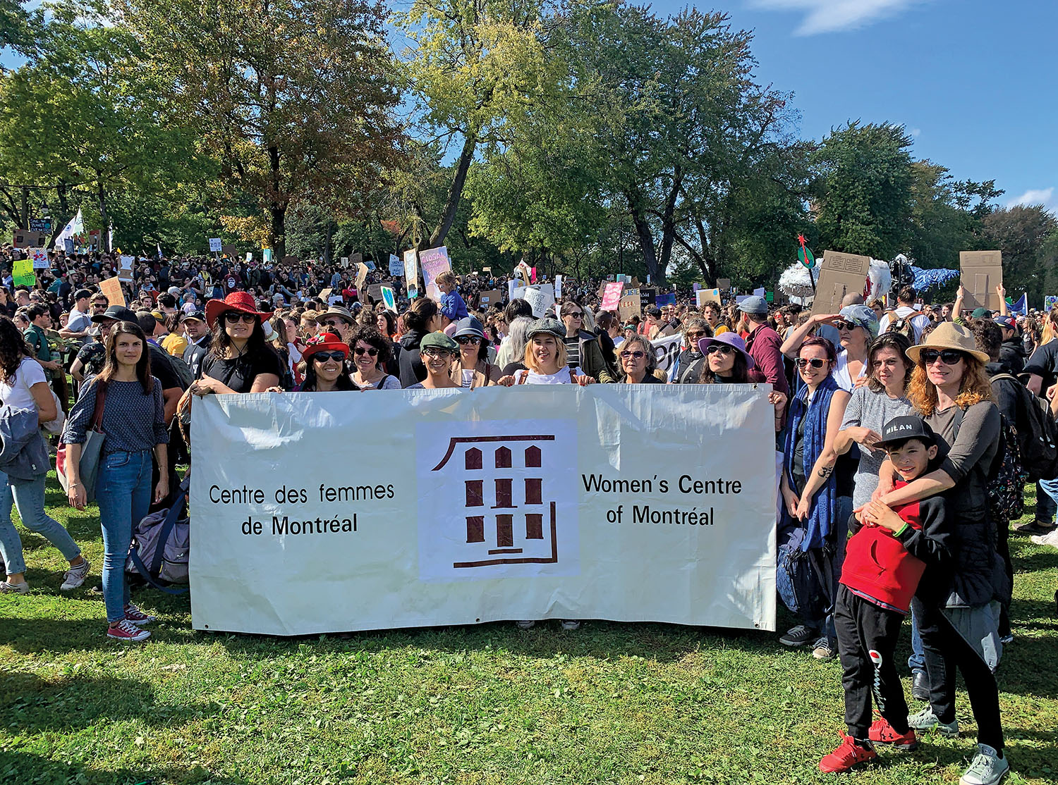 Photo couleur montrant une importante foule qui manifeste dans un parc. Au premier-plan, des femmes tiennent une banderole du Centre des femmes de Montréal.