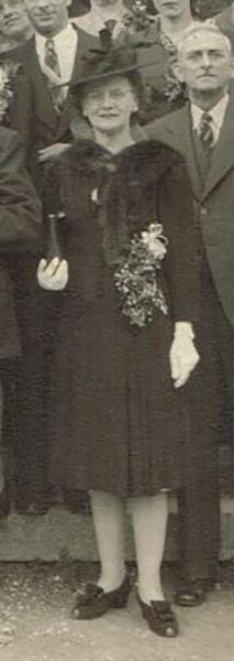 Photo noir et blanc recadrée autour d’une femme en format plein pied. Elle est entourée d’hommes et de femmes vêtues pour une occasion spéciale.