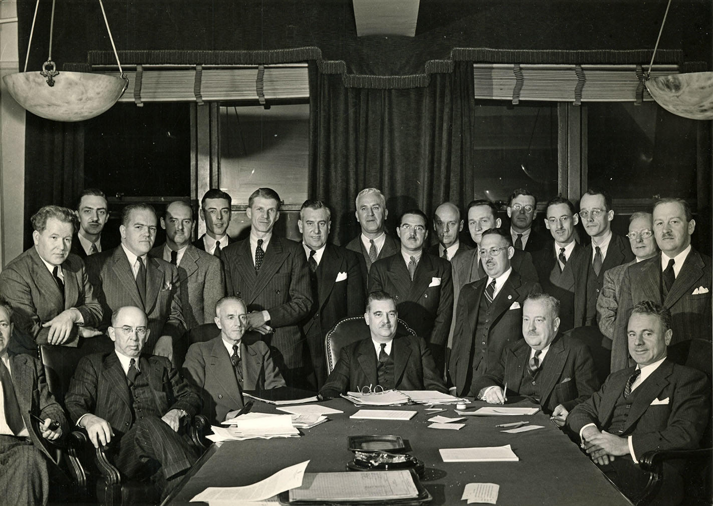 Photo en noir et blanc d’une assemblée d’hommes en complet prenant la pose derrière une table. Six sont assis et dix-sept sont debout.