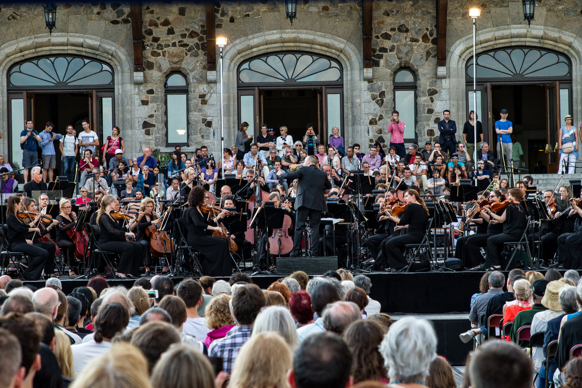 Un orchestre joue à l’extérieur, devant un édifice de pierre, et une foule assise écoute.