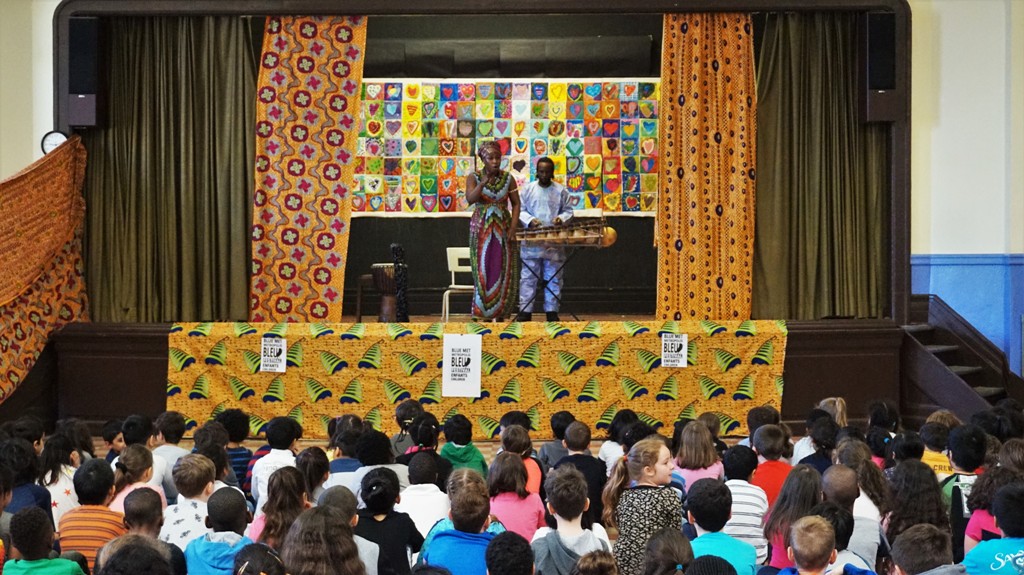 Spectacle de contes africains dans une école primaire. Deux artistes sont sur scène et les enfants sont assis par terre face à la scène.