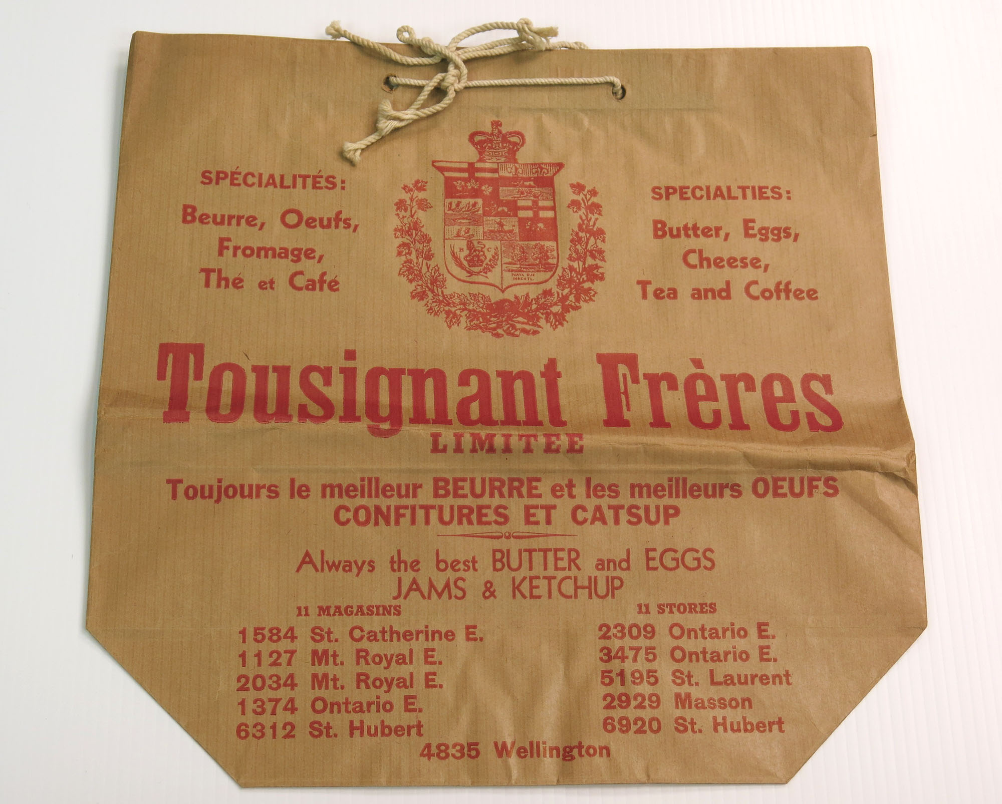 Sac en papier brun, avec des écritures rouges, de l’épicerie Tousignant & Frères. Des poignées en corde beige sont insérées dans le rebord en haut du sac.