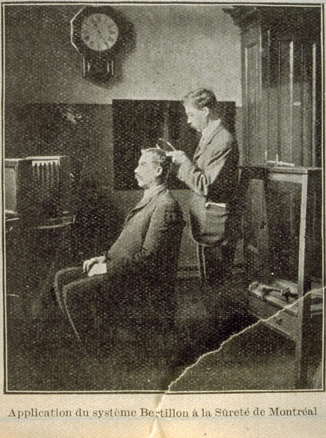 Coupure de journal en sépia qui montre une scène d’intérieur. Un homme debout mesure la tête d’un second homme assis sur une chaise, vue de profil.