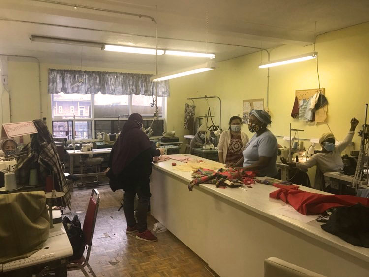 Cinq personnes sont dans un atelier de couture, deux assises devant des machines à coudre et trois debout autour d’une table haute.