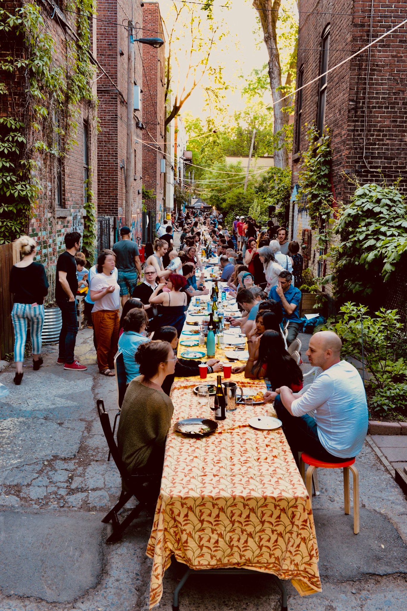 Photo couleur d’une longue tablée festive dans une ruelle remplie de convives assis et debout qui discutent, mangent et boivent.