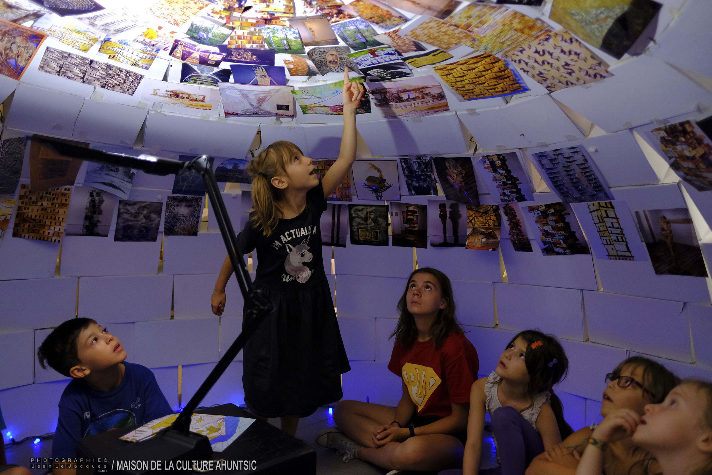 Six garçons et filles regardent des photos collées sur le plafond et les murs d’une petite maison.