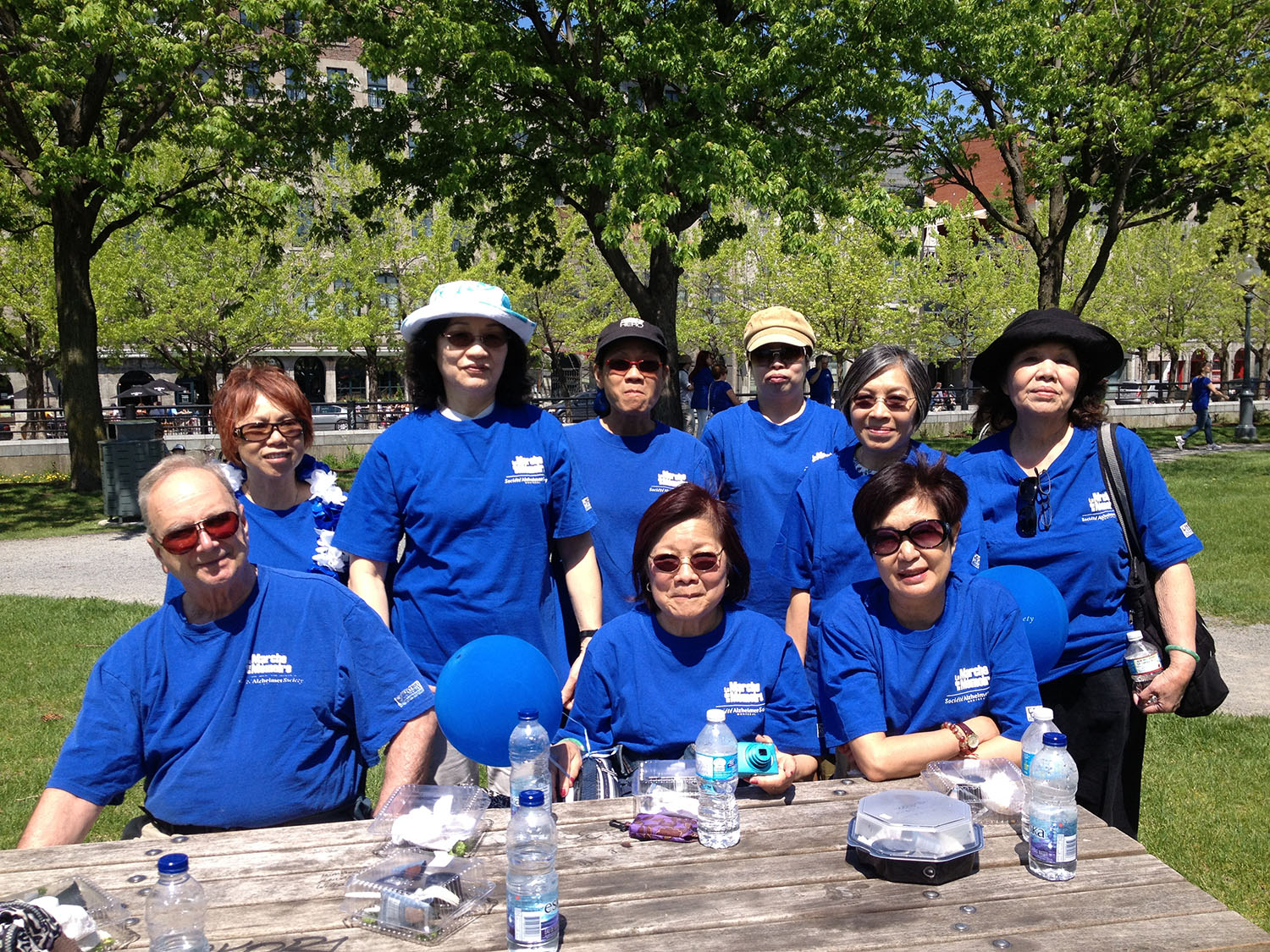 Neuf personnes portant un t-shirt bleu de la marche pour la Société Alzheimer prennent la pose et la pause dans un parc derrière une table de pique-nique. 