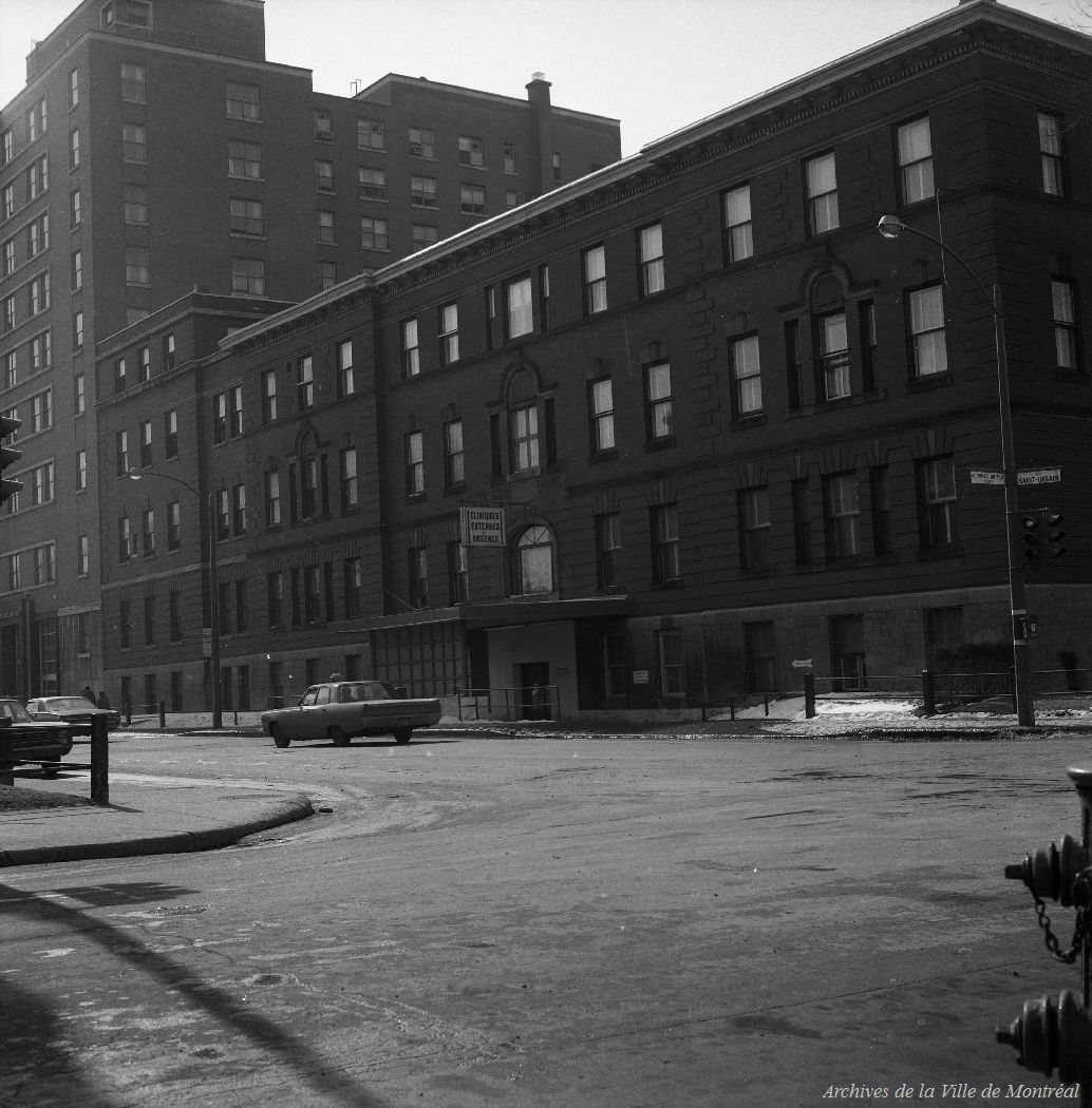 Photographie en noir et blanc montrant un bâtiment de briques au coin de deux rues. Un panneau fixé sur la façade porte l’inscription « Cliniques externes et urgence ». 