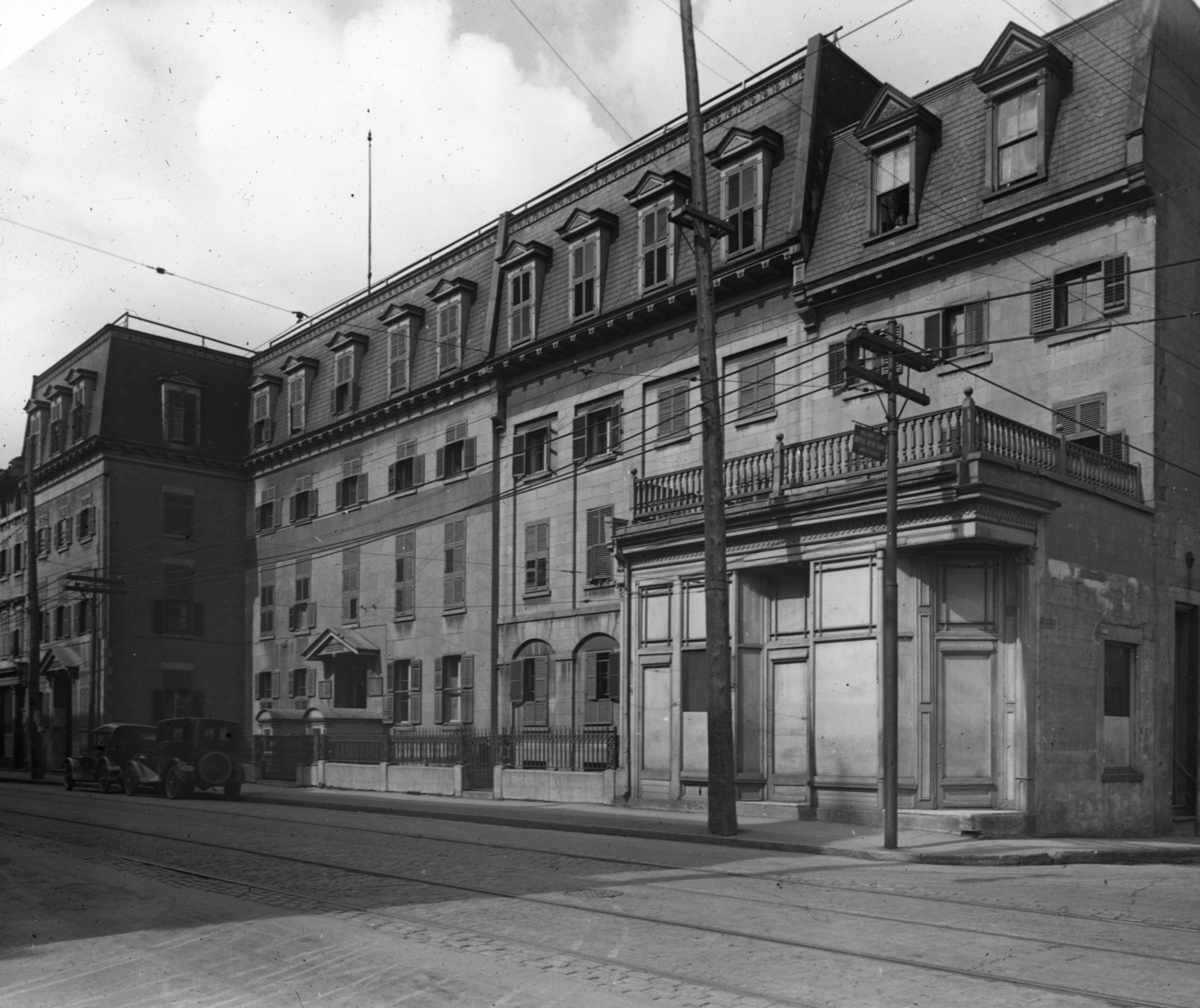 Photographie de la façade de l'hôpital Notre-Dame lorsqu'il était situé sur la rue Notre-Dame. On y remarque des poteaux électriques, les câbles servant à la circulation du tramway et deux voitures arrêtées devant l'hôpital.