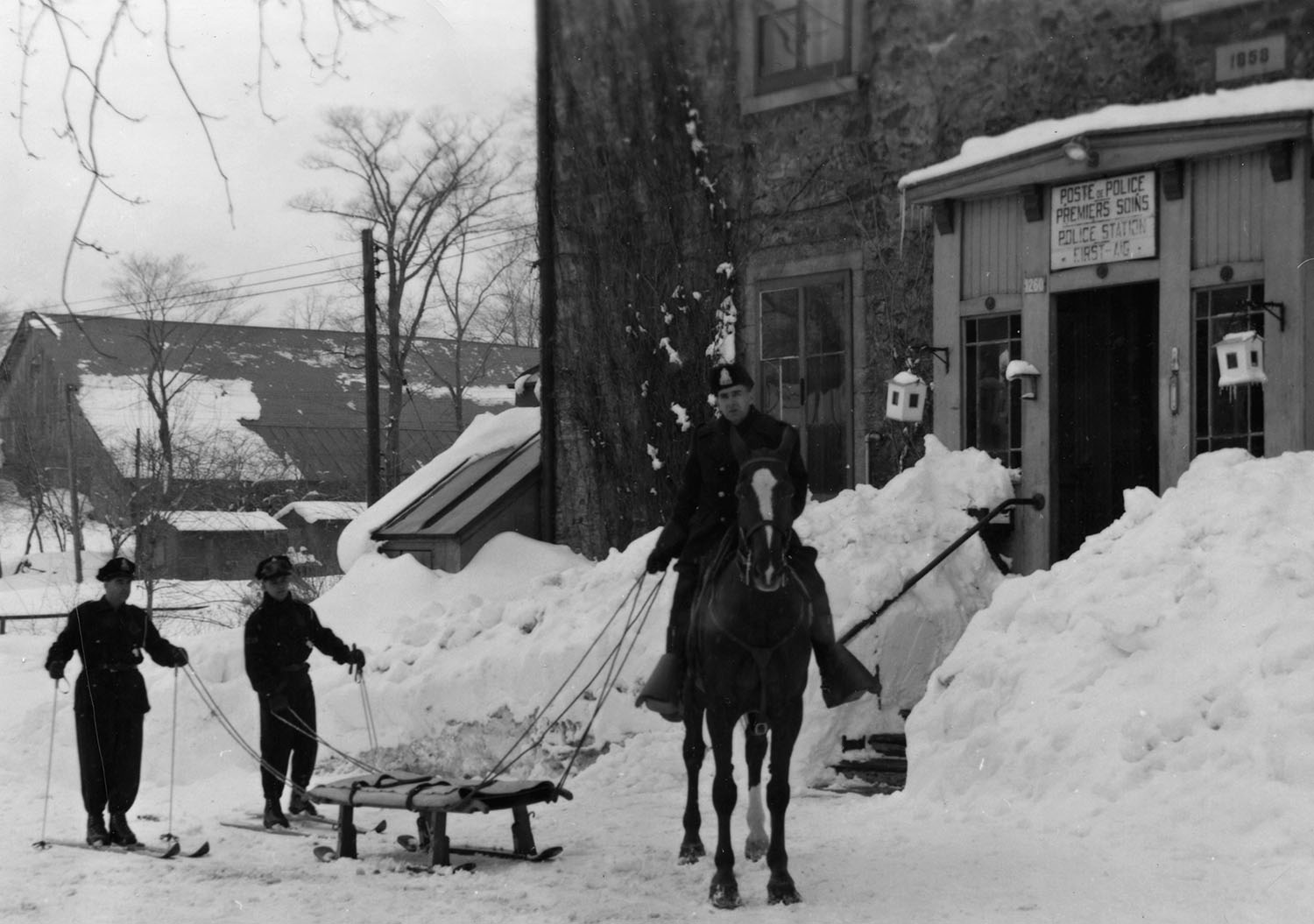 Photographie en noir et blanc sur laquelle on voit un policier à cheval tirant une civière attelée à l’animal. Derrière la civière, il y a deux policiers à ski.