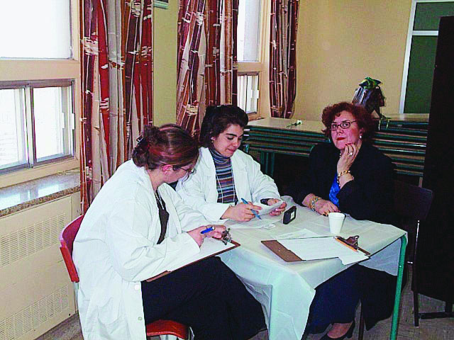 Trois femmes sont assises autour d’une petite table carrée : les deux jeunes filles portent un sarrau blanc et prennent des notes en écoutant la dame.