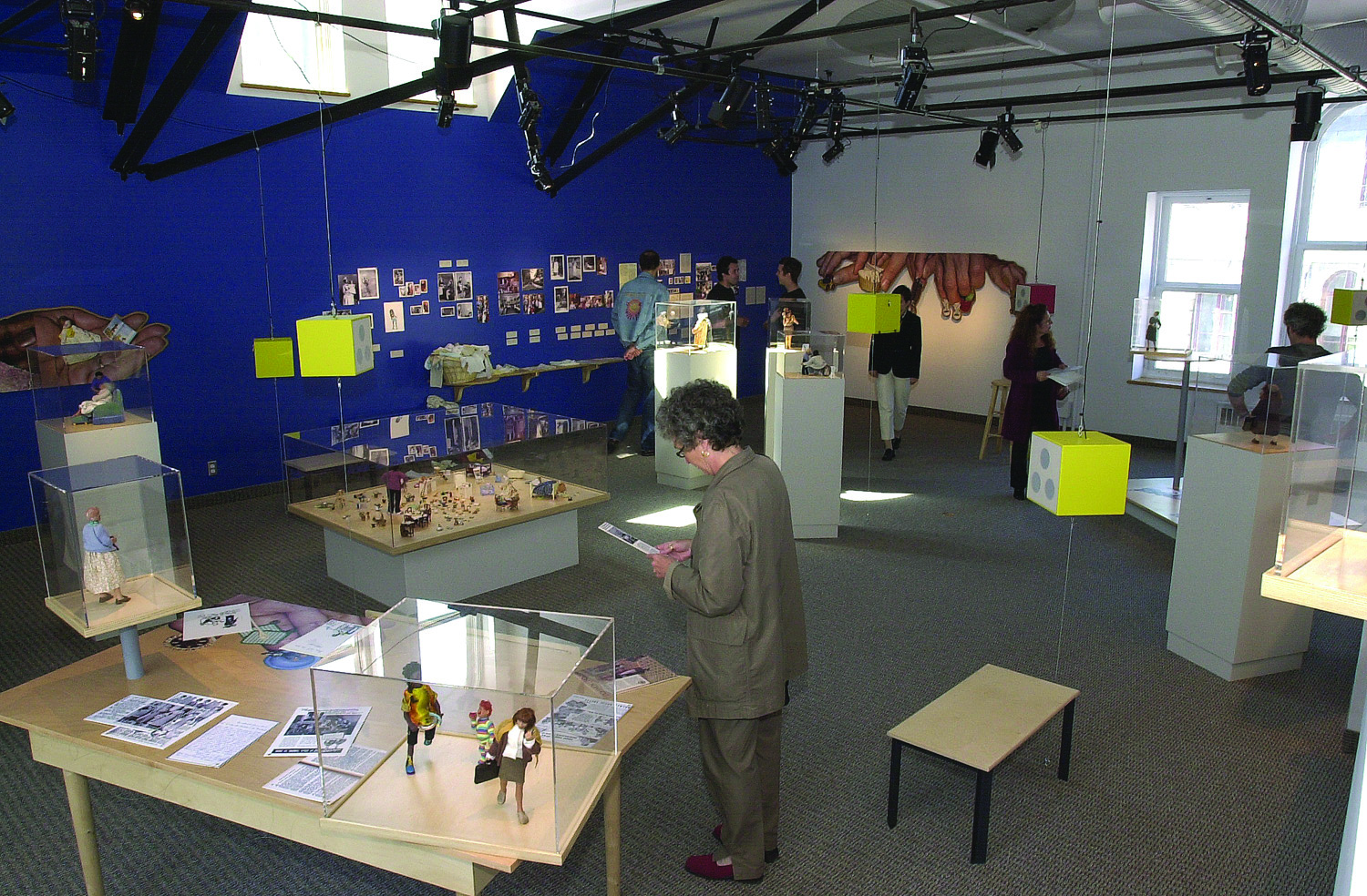 Vue d’ensemble d’une salle d’exposition avec des petits personnages dans des vitrines, des boîtes jaunes suspendues pour écouter de l’audio et des photos au mur. Sept personnes visitent l’exposition.