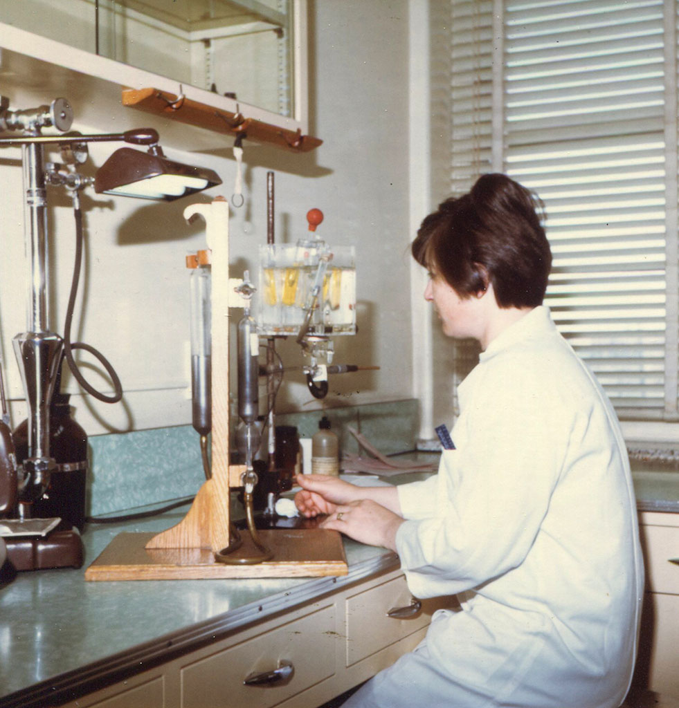 Photo couleur d’une jeune femme travaillant dans un laboratoire. On la voit de côté, elle manipule un instrument sur un comptoir devant elle. 
