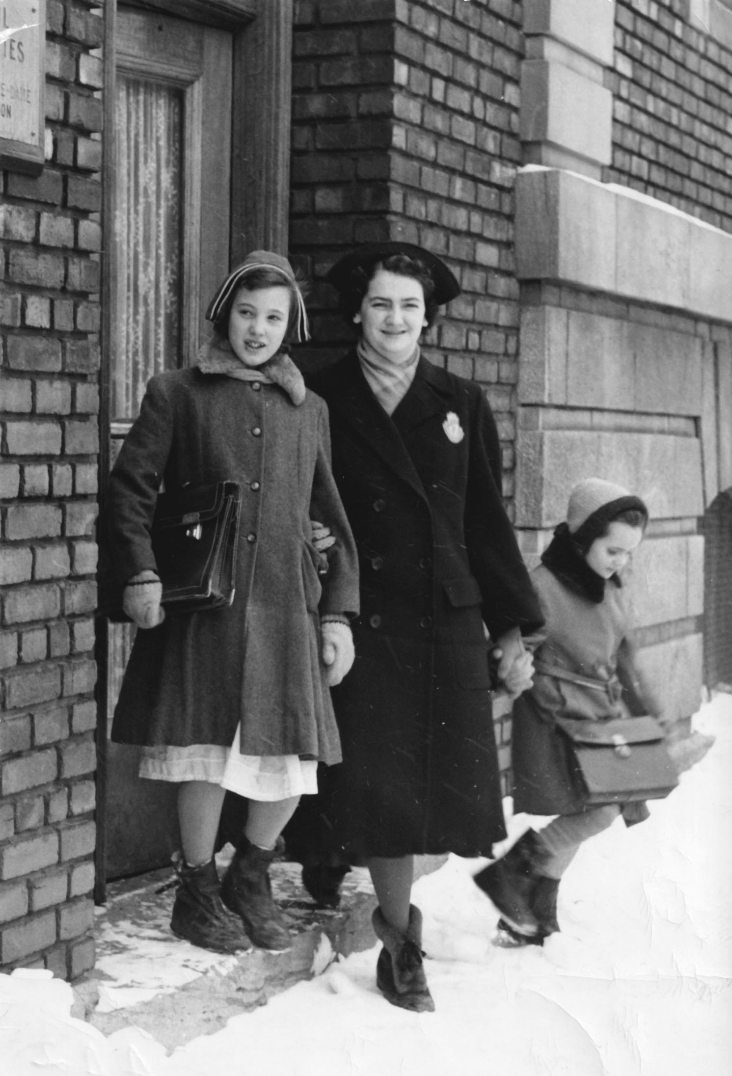Une femme habillée avec un manteau et un chapeau d’hiver tient, de chaque main, une fillette. Elles sont devant une porte d’entrée. 
