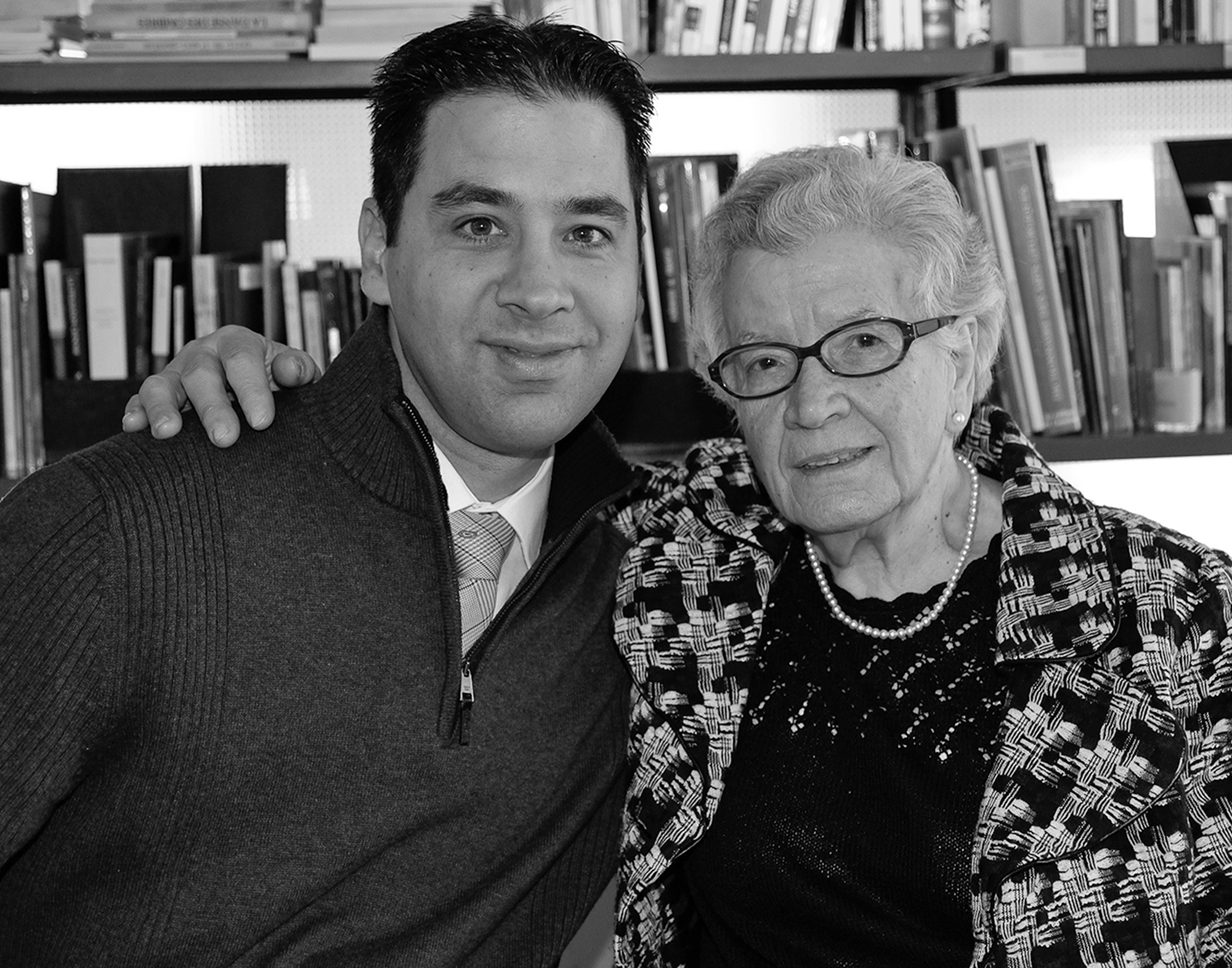 Photo en noir et blanc d’une jeune homme et de sa grand-mère se tenant par les épaules, collés l’un contre l’autre. Une bibliothèque en arrière-plan.   