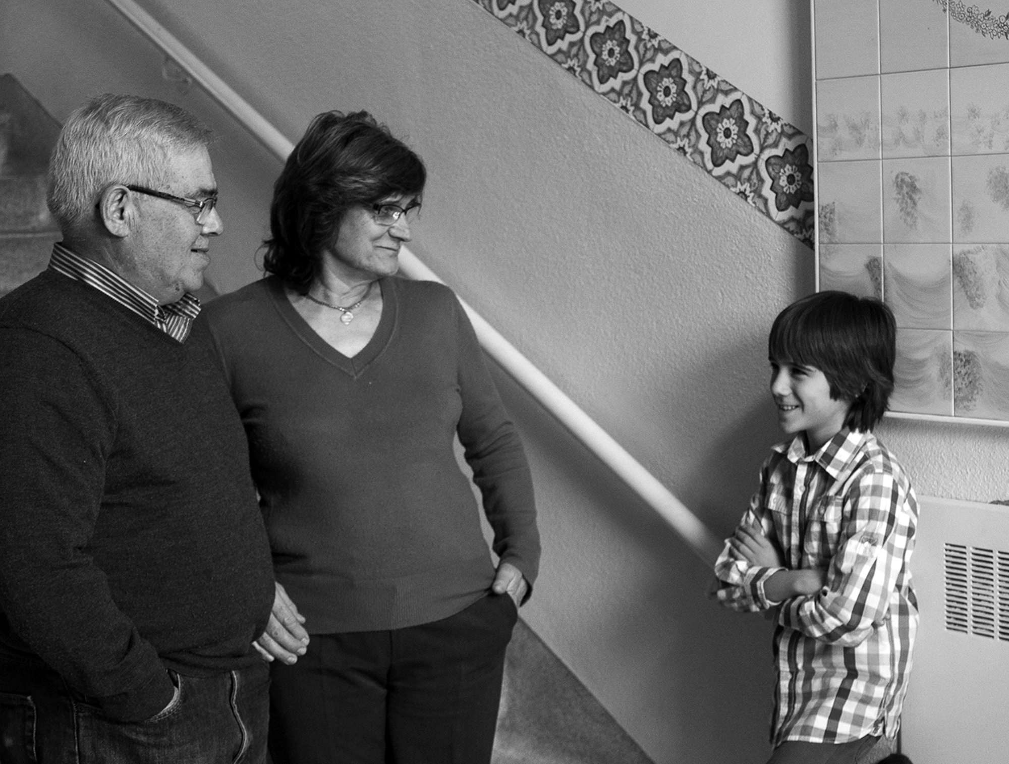 Photo en noir et blanc d’un jeune garçon, debout, regardant son grand-père et sa grand-mère, aussi debout. Ils sourient tous les trois.   