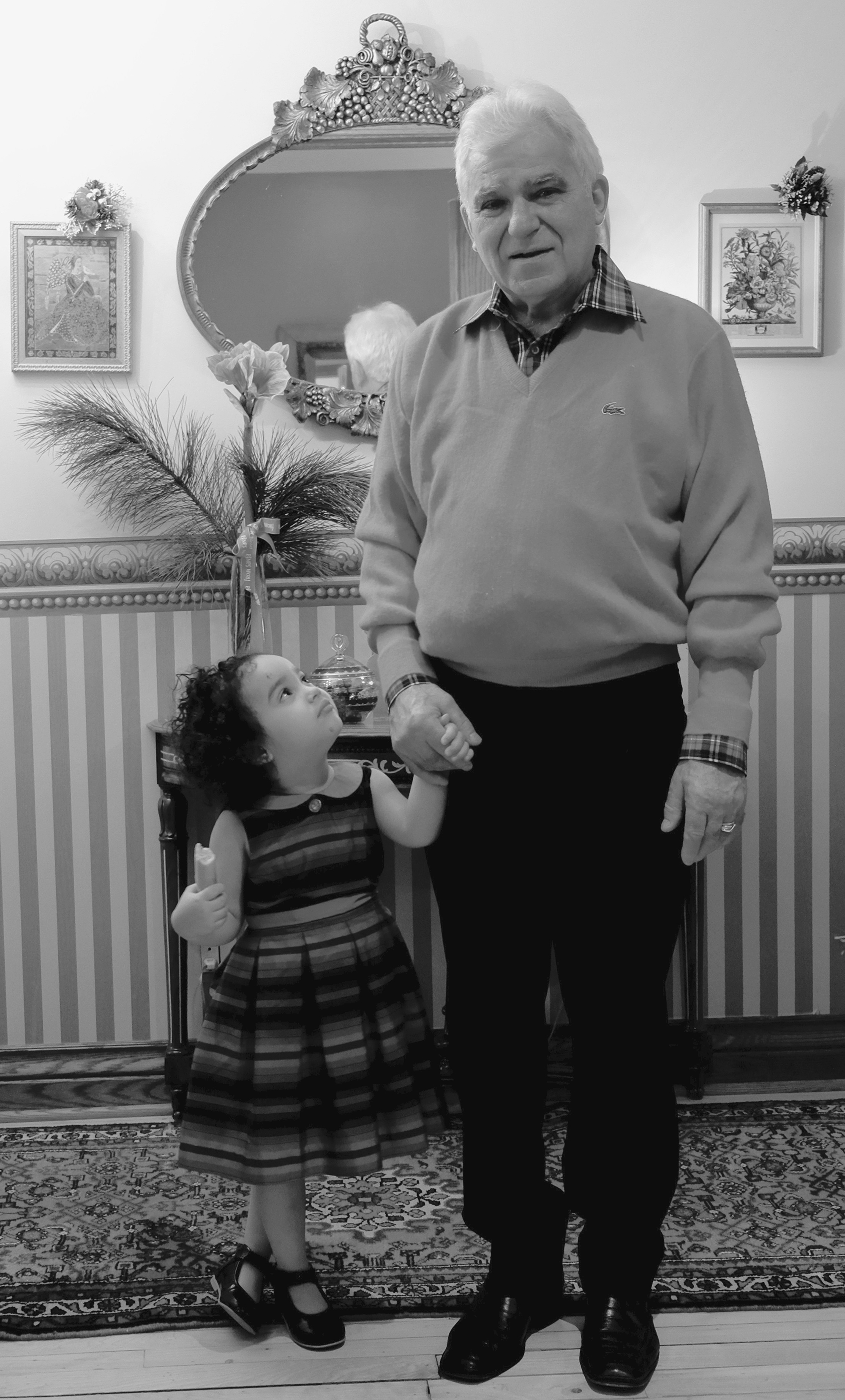 Photo en noir et blanc montrant une petite-fille, debout, qui lève les yeux vers son grand-père, debout à côté d’elle et lui tenant la main. .