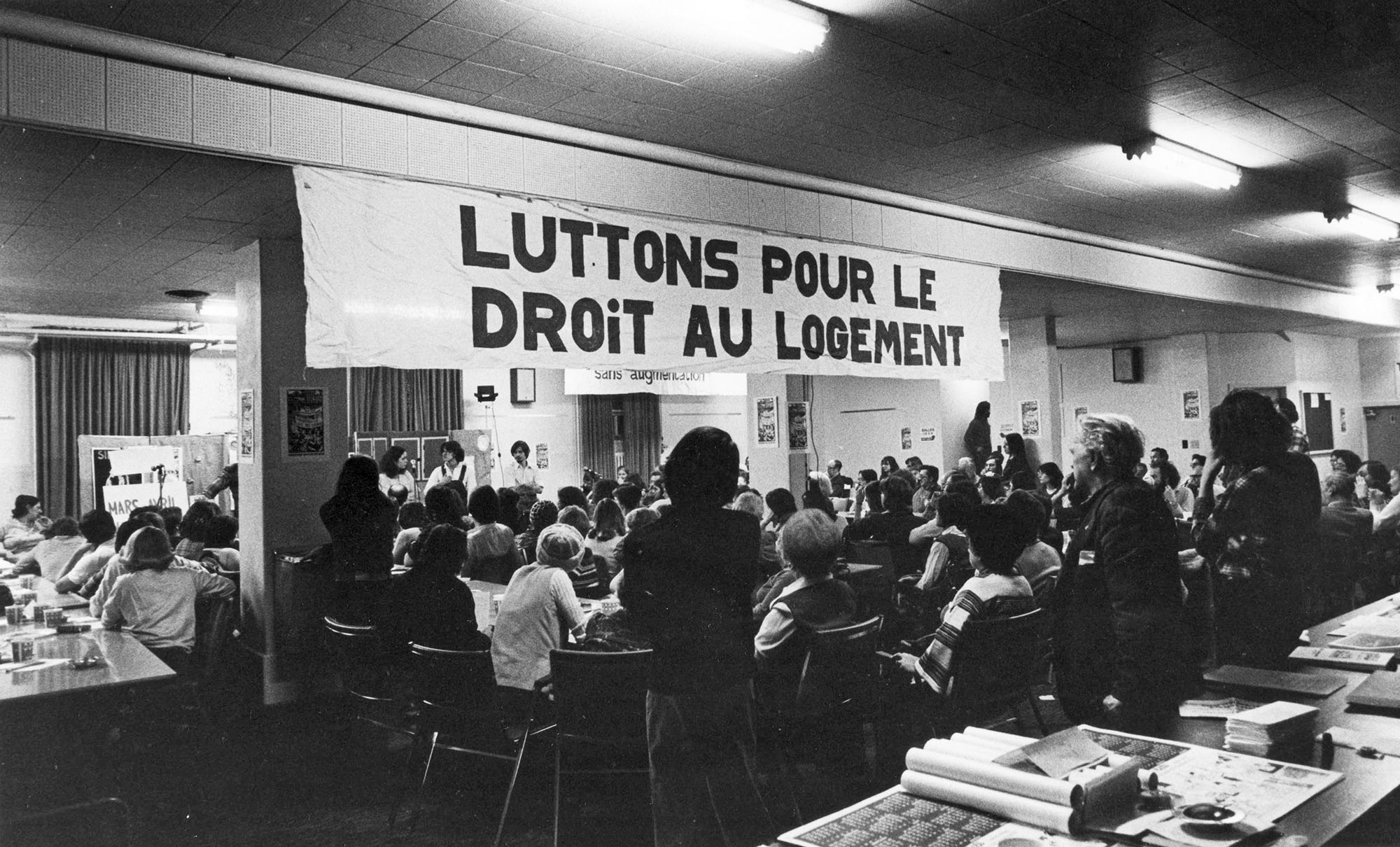 Une cinquantaine de personnes sont assises ou debout, de dos, dans une salle. Une affiche, sur laquelle on peut lire « Luttons pour le droit au logement », est suspendue au plafond.