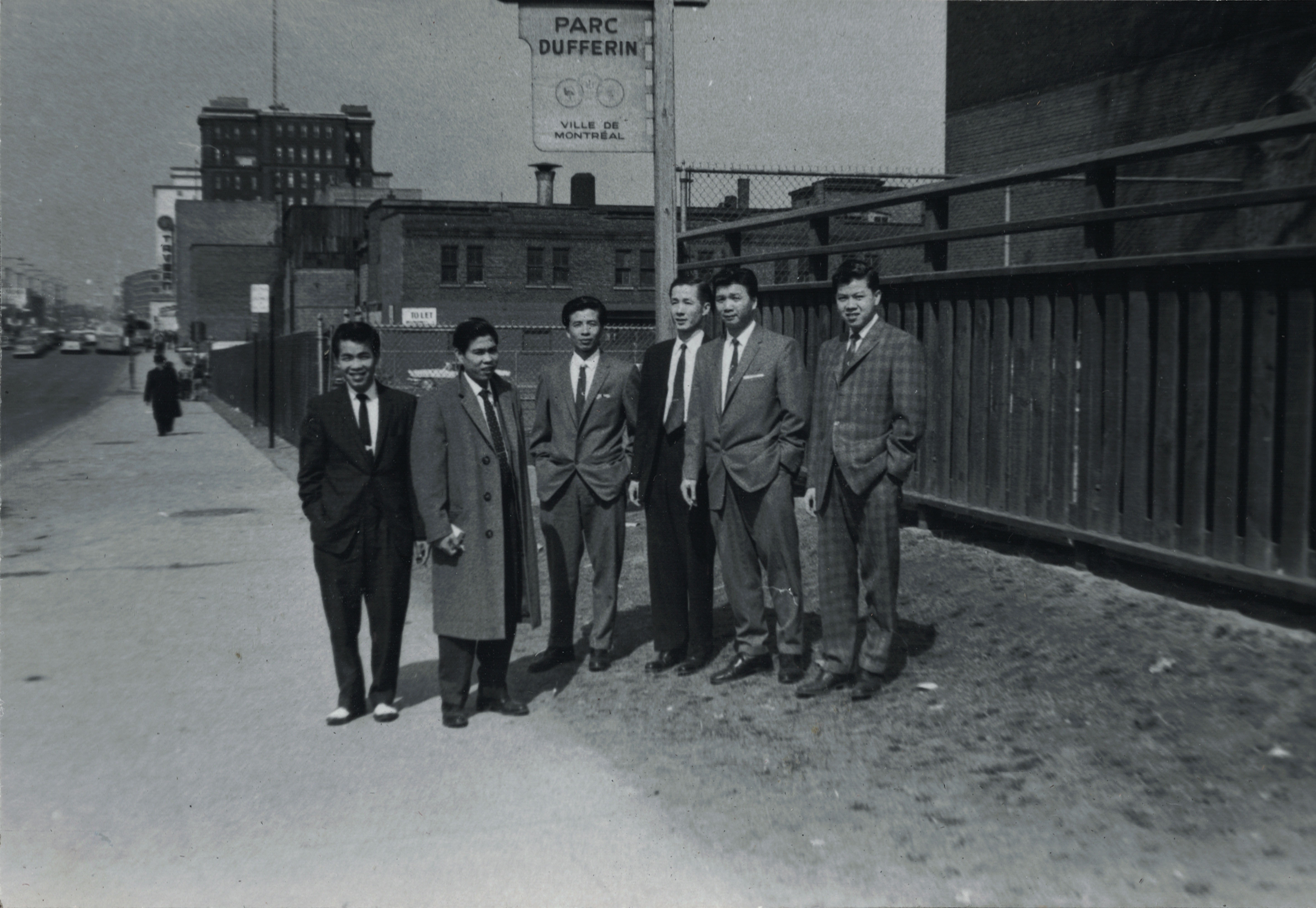 Photo en noir et blanc montrant six hommes d’origine chinoise se tenant en partie sur le trottoir et en partie sur la pelouse devant une pancarte du parc Dufferin. 