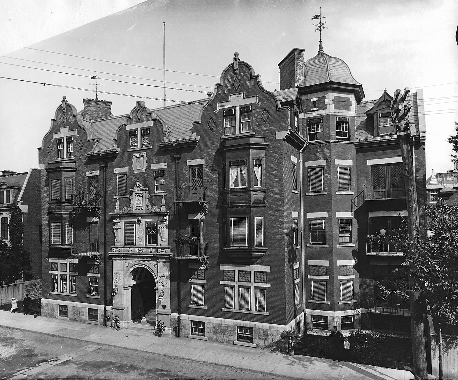 Photographie en noir et blanc de la façade de l’édifice Marlborough. Des girouettes sont visibles sur le sommet des toitures des tourelles à gauche et à droite, et des fils électriques passent au-dessus de l’édifice, au centre.