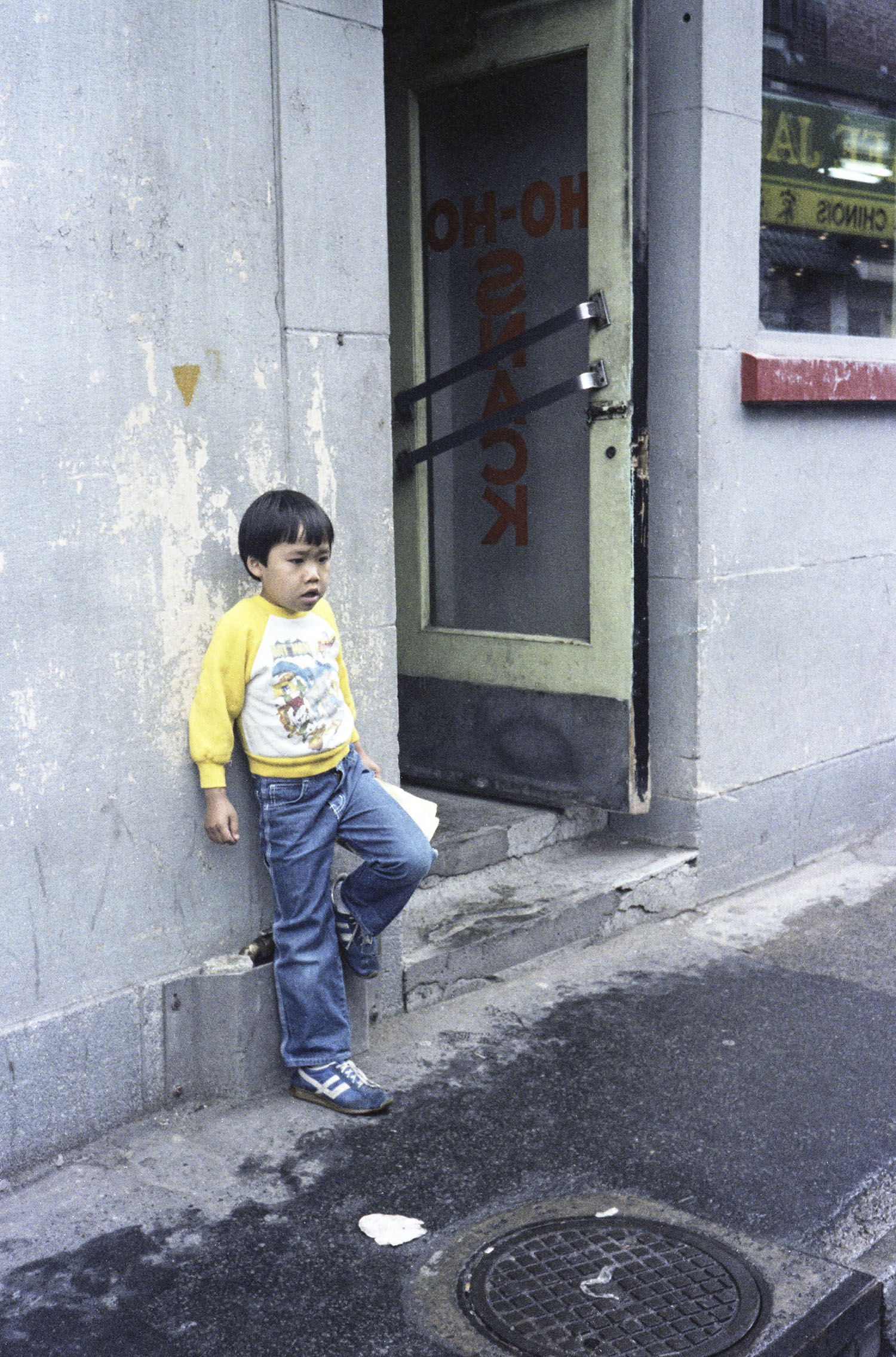 Un jeune enfant d’origine chinoise se tient debout contre le mur à côté de la porte d’entrée d’un restaurant. On peut lire Ho Ho Snack à l’envers sur la porte vitrée.