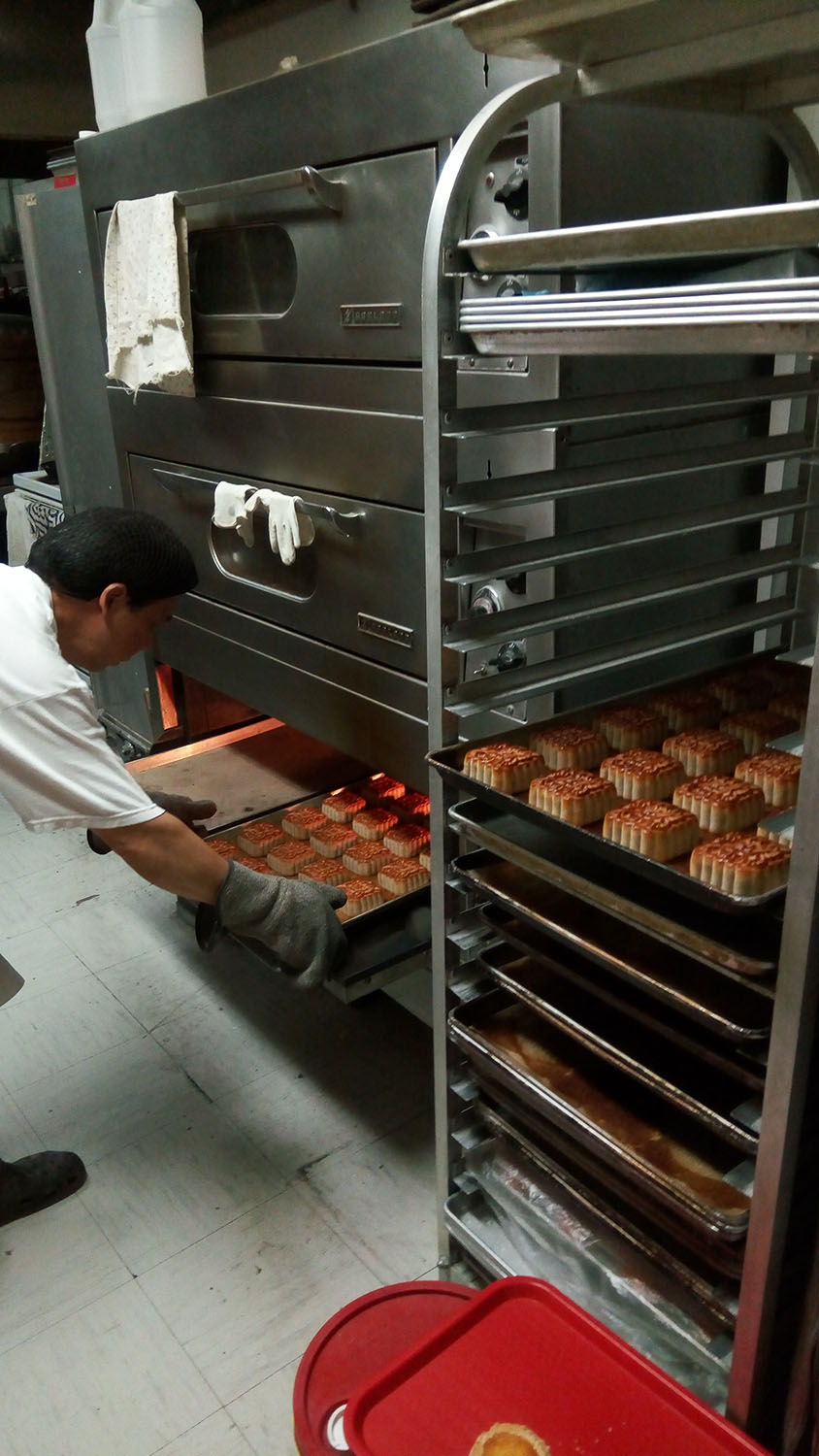 A man bends over to put a sheet of mooncakes into the bottom section of a large triple-deck oven. 