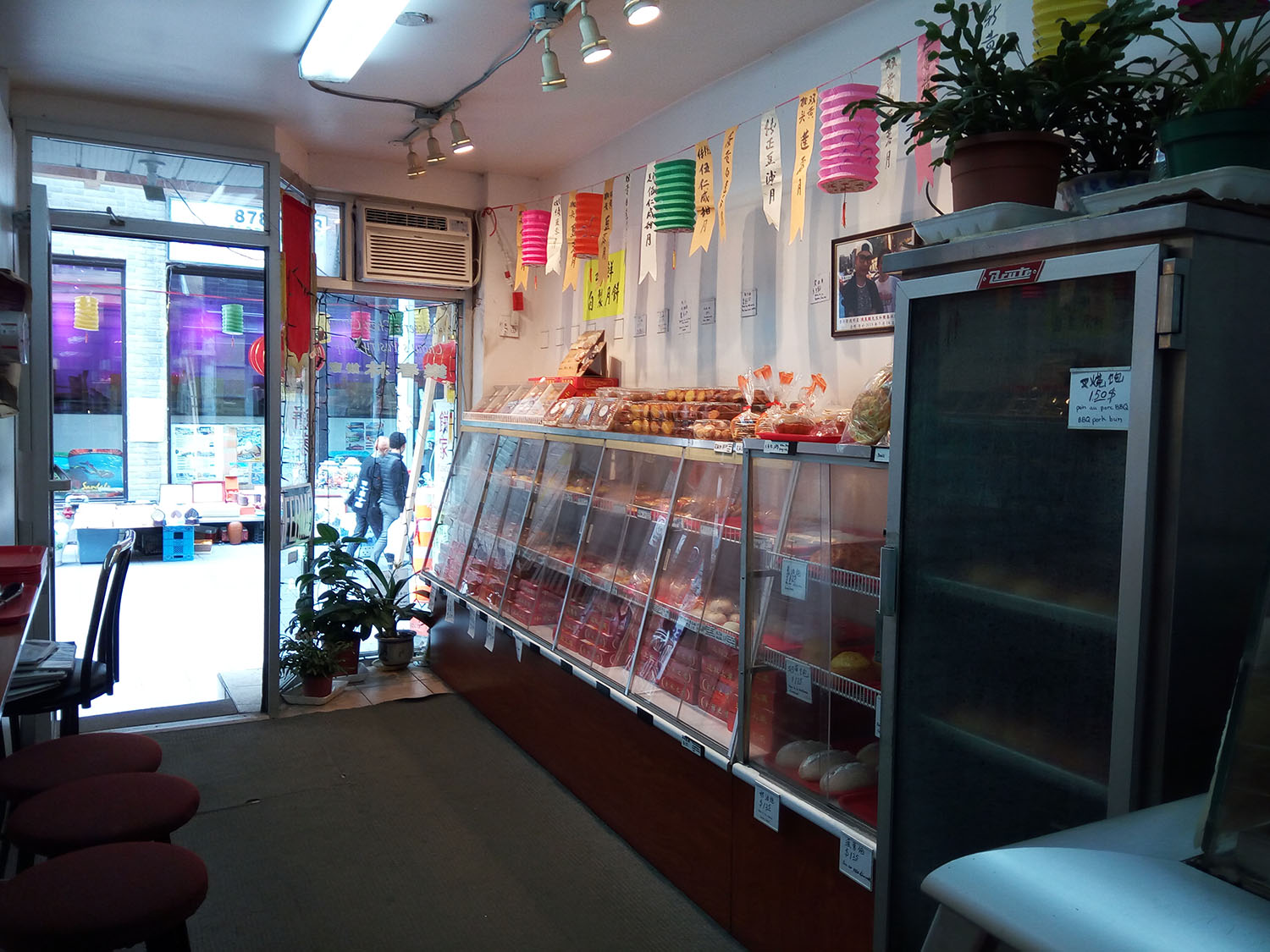 Colour photo of the interior of a traditional Chinese pastry shop. The colour photograph shows display cases on the right, seating on the left, and, in the background, a glass door opening onto rue De La Gauchetière. 
