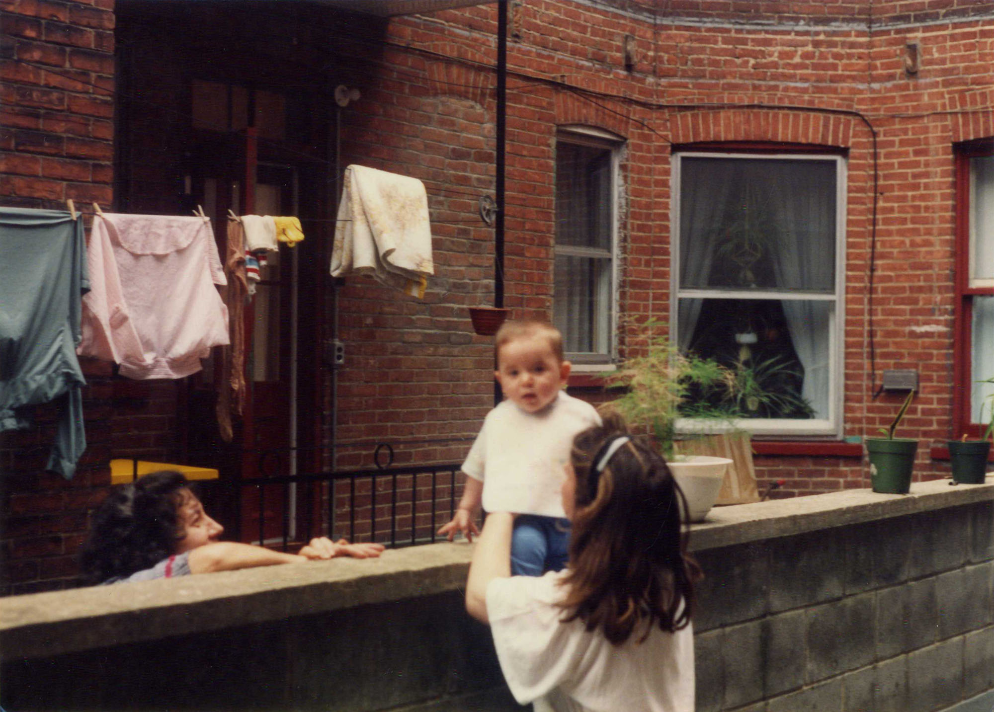 Une femme, de dos, tient son bébé qui est assis sur un muret dans la cour derrière le triplex familial. Une voisine le regarde. 