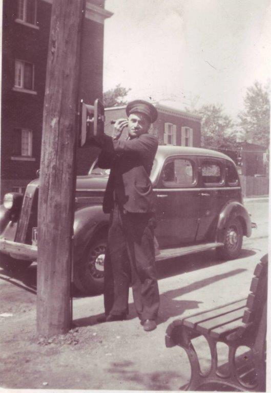 Photo noir et blanc d’un chauffeur de taxi prenant un appel d’une boîte téléphonique accrochée à un poteau sur un trottoir, sa voiture est stationnée derrière lui sur une rue résidentielle.