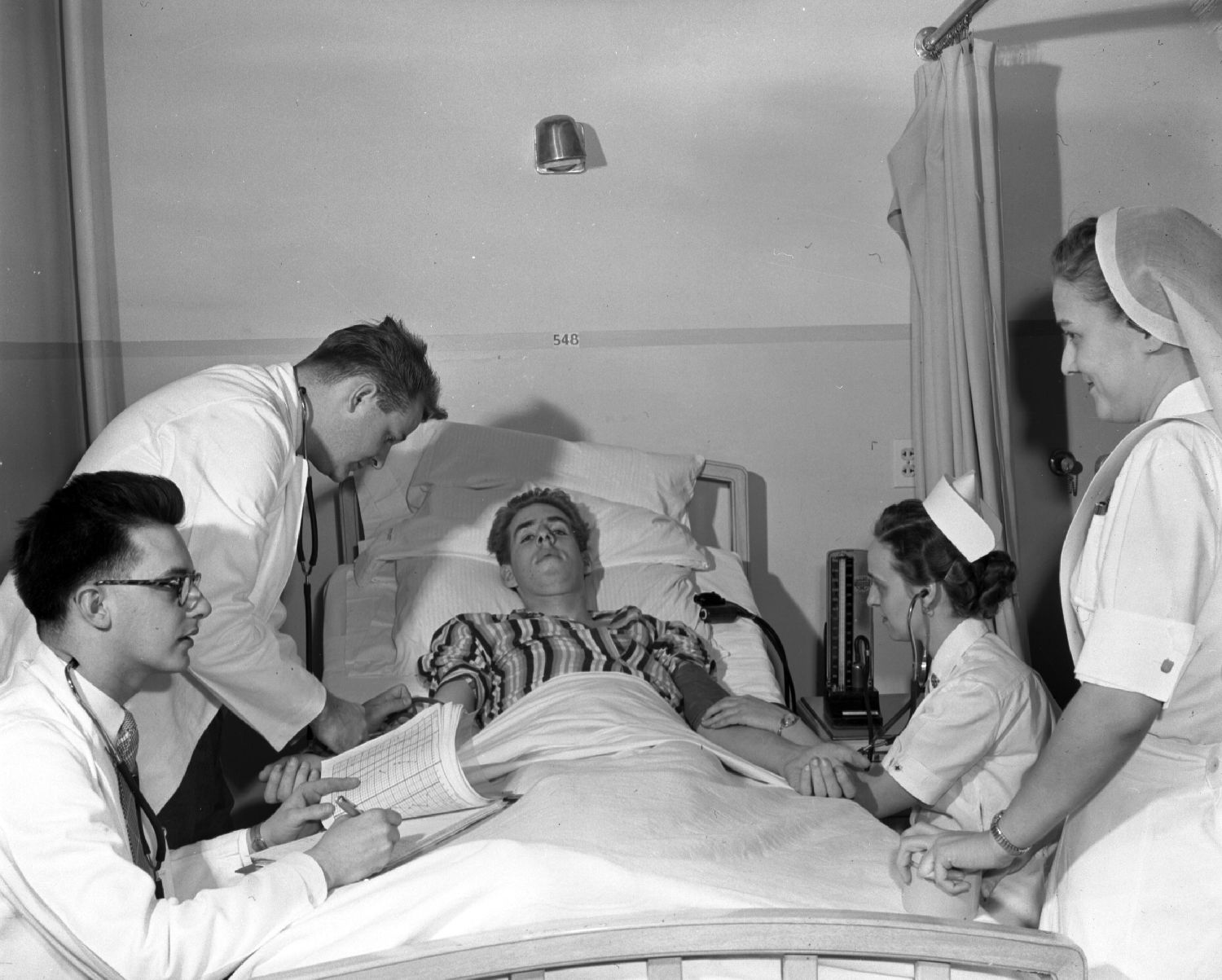 Photo en noir et blanc montrant un malade alité. D’un côté se trouvent deux infirmières et de l’autre deux médecins.