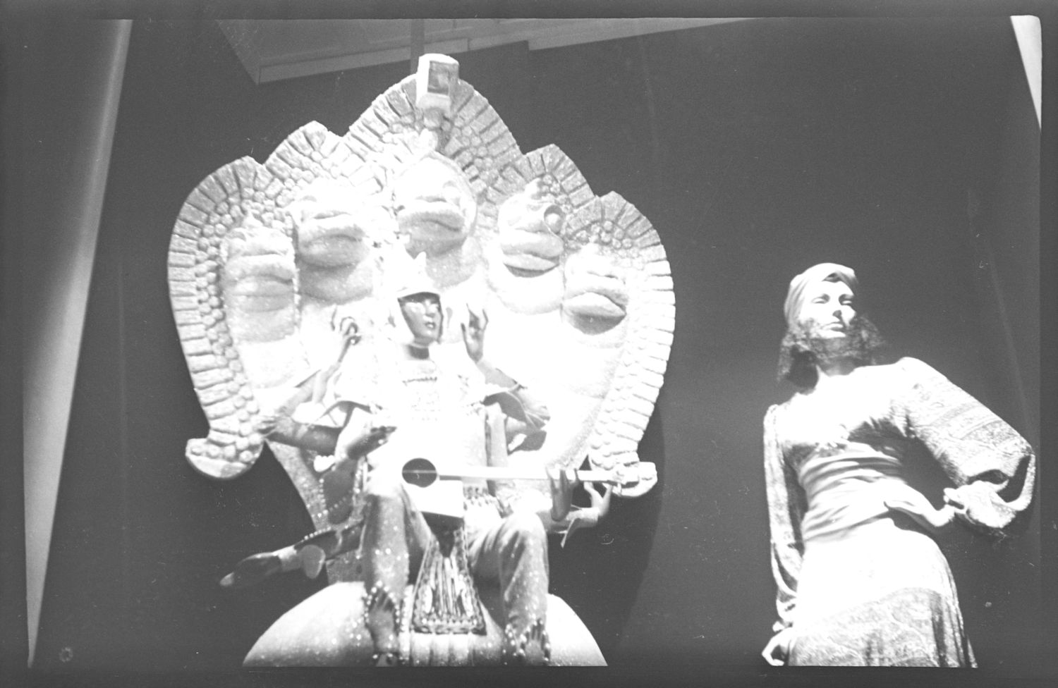 Photo en noir et blanc d’une vitrine de grand magasin en 1941. On voit un mannequin féminin portant une robe et un turban. Près d’elle, assis sur un imposant trône d’allure orientale, se trouve un musicien tibétain jouant d’un instrument traditionnel. 