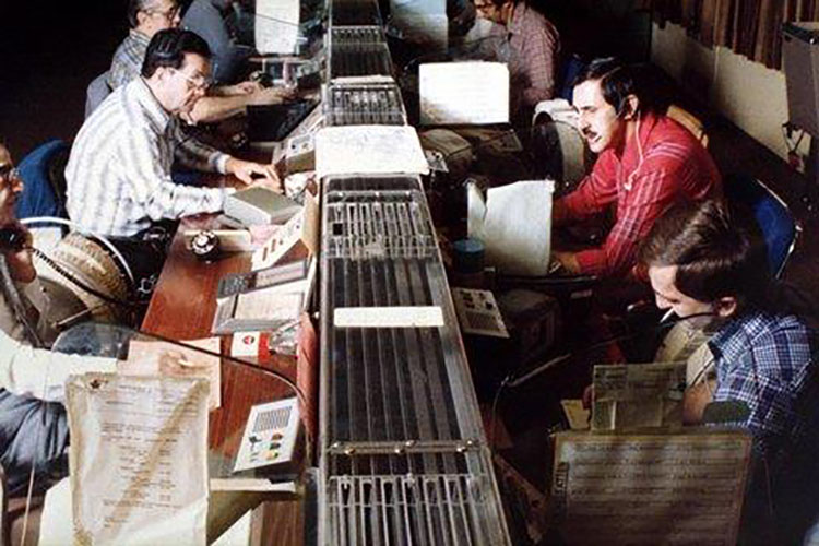 Photo couleur d’un centre de télécommunications avec une longue série de tables avec des appareils au centre. Des hommes sont assis de chaque côté.