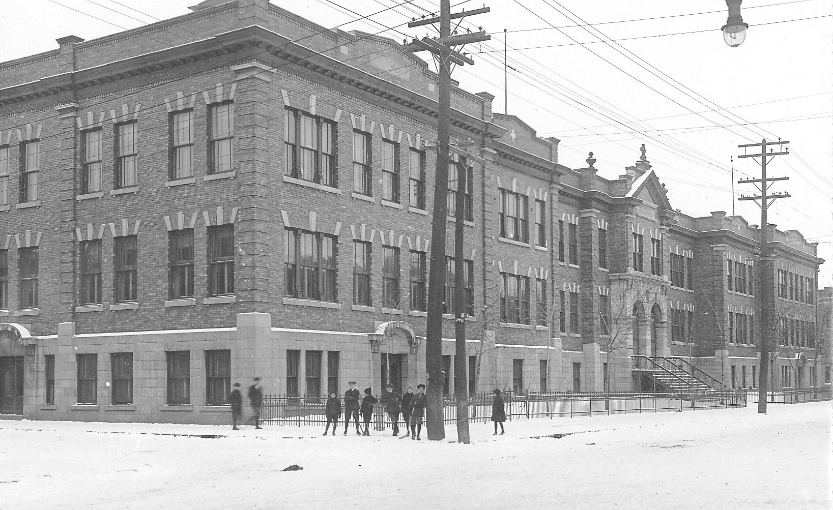 Photo noir et blanc de la façade et d’un côté d’un grand édifice à trois  étages. Neuf personnes sont debout au coin de la rue devant l’édifice. 