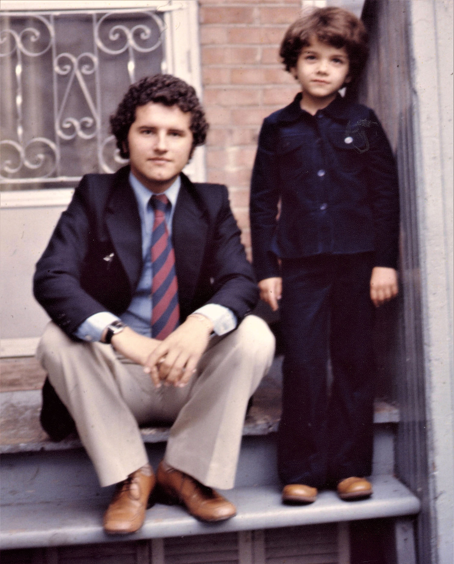 Photo couleur d’un jeune homme assis dans les marches de la maison familiale et son jeune frère debout à ses côtés. 