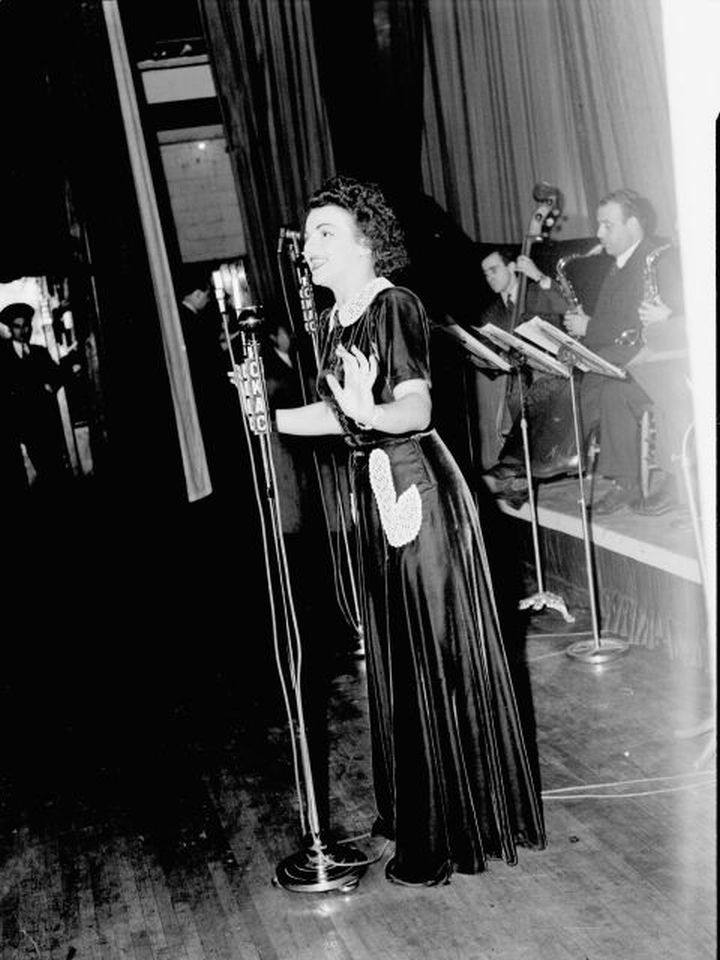 La chanteuse Alys Roby accompagnée par un orchestre sur la scène d'une salle de spectacle lors de la radiodiffusion de l'émission "Tourbillon de la Gaieté" de l'auteur Roger Marien par la station C.K.A.C. à Montréal.