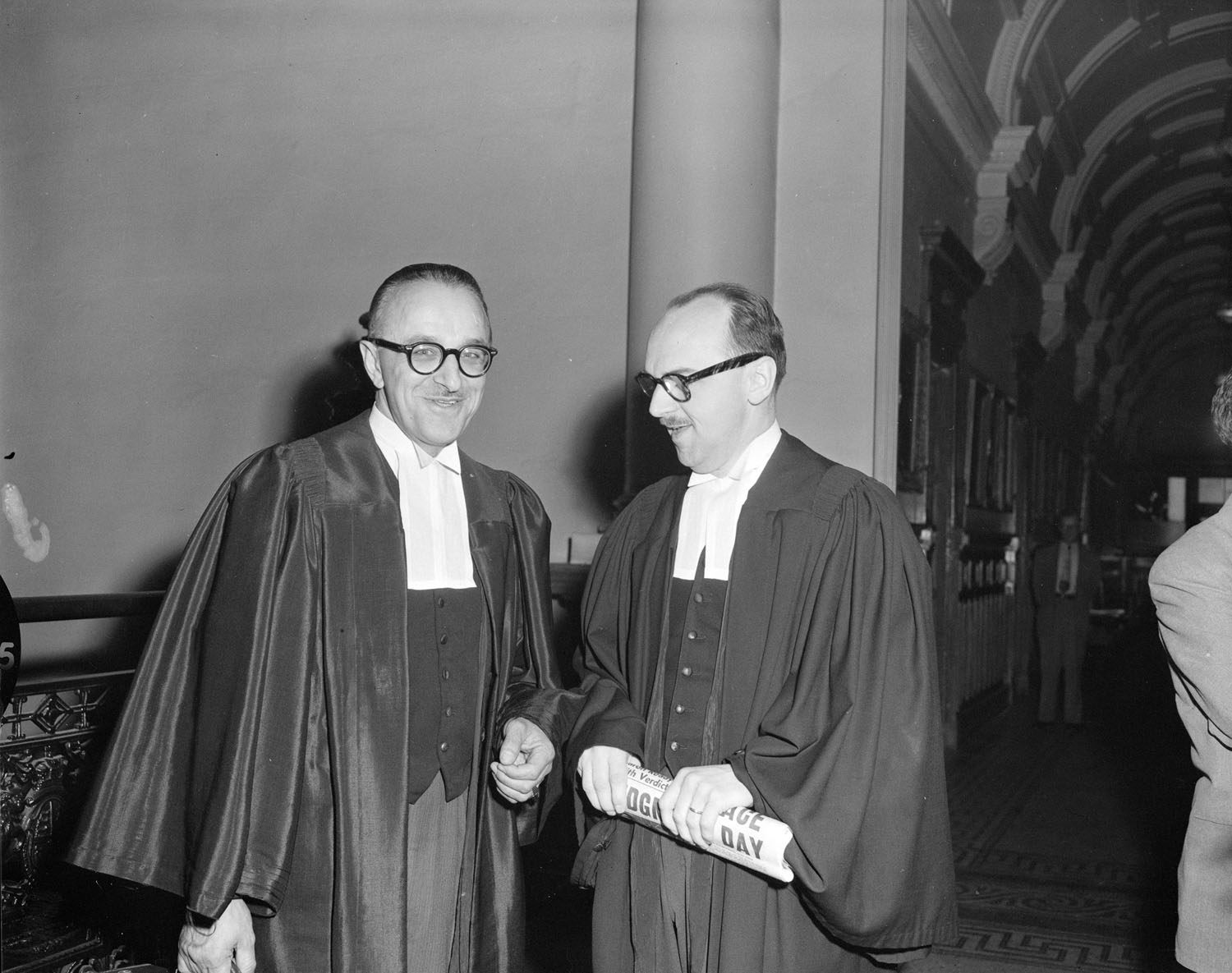 Deux hommes à lunettes portant la toge d’avocat se tiennent debout dans une salle non identifiée. L’homme de droite tient un journal dans ses mains.
