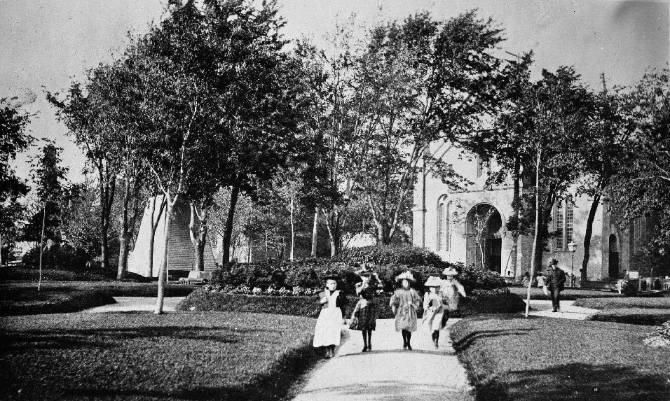 Photographie en noir et blanc représentant un groupe d’enfants marchant dans un parc, avec à l’arrière-plan une église.