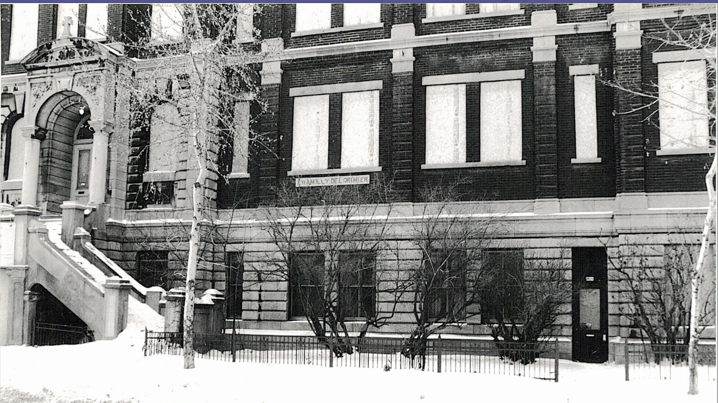 Photo noir et blanc montrant une partie de la façade d’un édifice ancien ayant abrité une école. On peut y lire l’inscription « Chamilly-de-Lorimier ». Les fenêtres des étages sont placardées.  