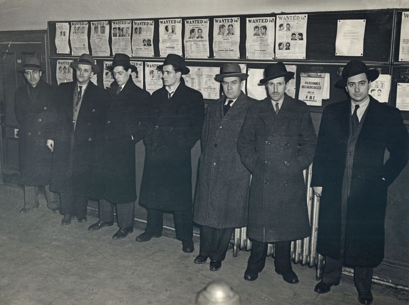 Photo noir et blanc de sept enquêteurs de police devant un babillard de personnes recherchées.