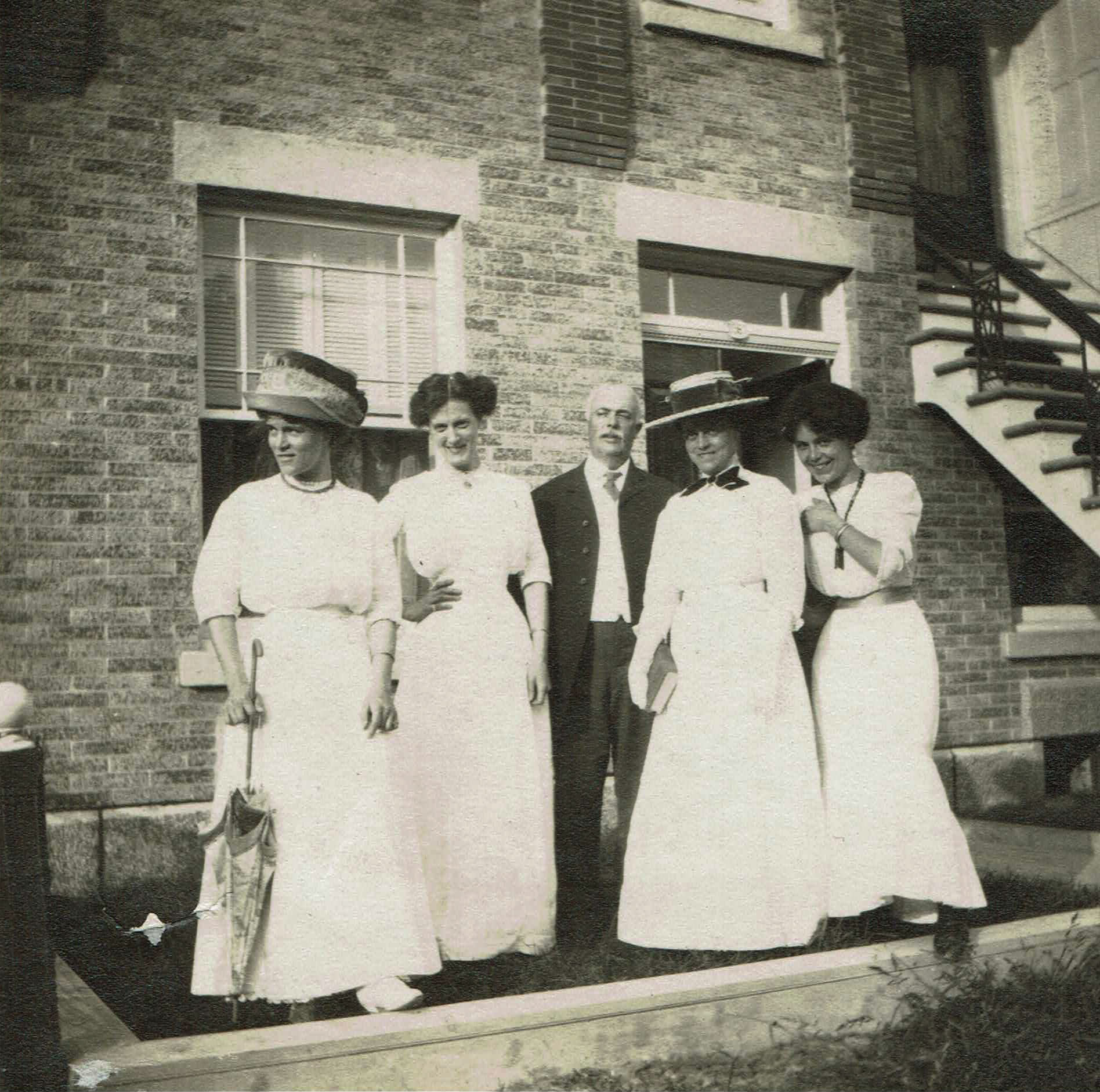 Photographie en noir et blanc représentant un homme d’âge mûr entouré de quatre jeunes femmes devant un immeuble de brique.