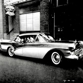 Photo en noir et blanc d'une voiture devant un édifice avec l'enseigne de Wing's Noodle. Une femme et quatre enfants sont dans la voiture aux fenêtres ouvertes.