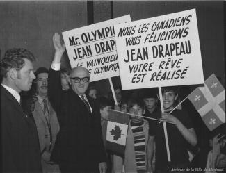 Photographie du maire saluant au centre, entouré d'adultes et de jeunes. Une pancarte affiche « Nous les Canadiens vous félicitons Jean Drapeau votre rêve est réalisé ».