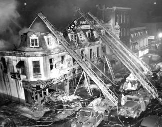 Photographie en noir et blanc montrant les décombres d’un édifice après un incendie. Des camions de pompier sont visibles sur la droite de la photographie.