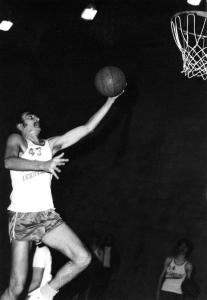Photo en noir et blanc d’un homme jouant au basketball. Il est dans les airs avec le ballon dans une main en direction du panier à la droite de la photo.